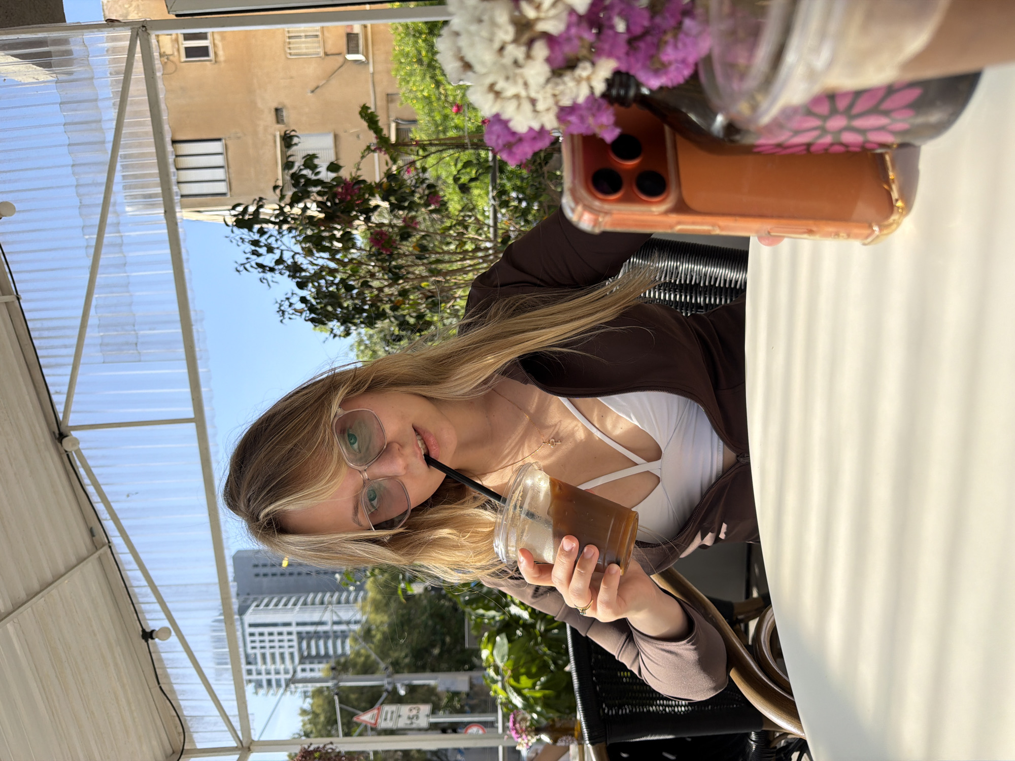 A blonde woman wearing glasses, a white top, and a brown jacket is seen drinking from a plastic cup with a straw at an outdoor café. She is holding the cup with her right hand and has a ring on her ring finger. An orange smartphone with a floral-patterned case rests on the table. A bouquet of white and purple flowers is near the phone. The café appears to be under a translucent roof. The background includes a building with windows, trees, and another taller building. The city of Bat Yam, Israel is visible in the background.