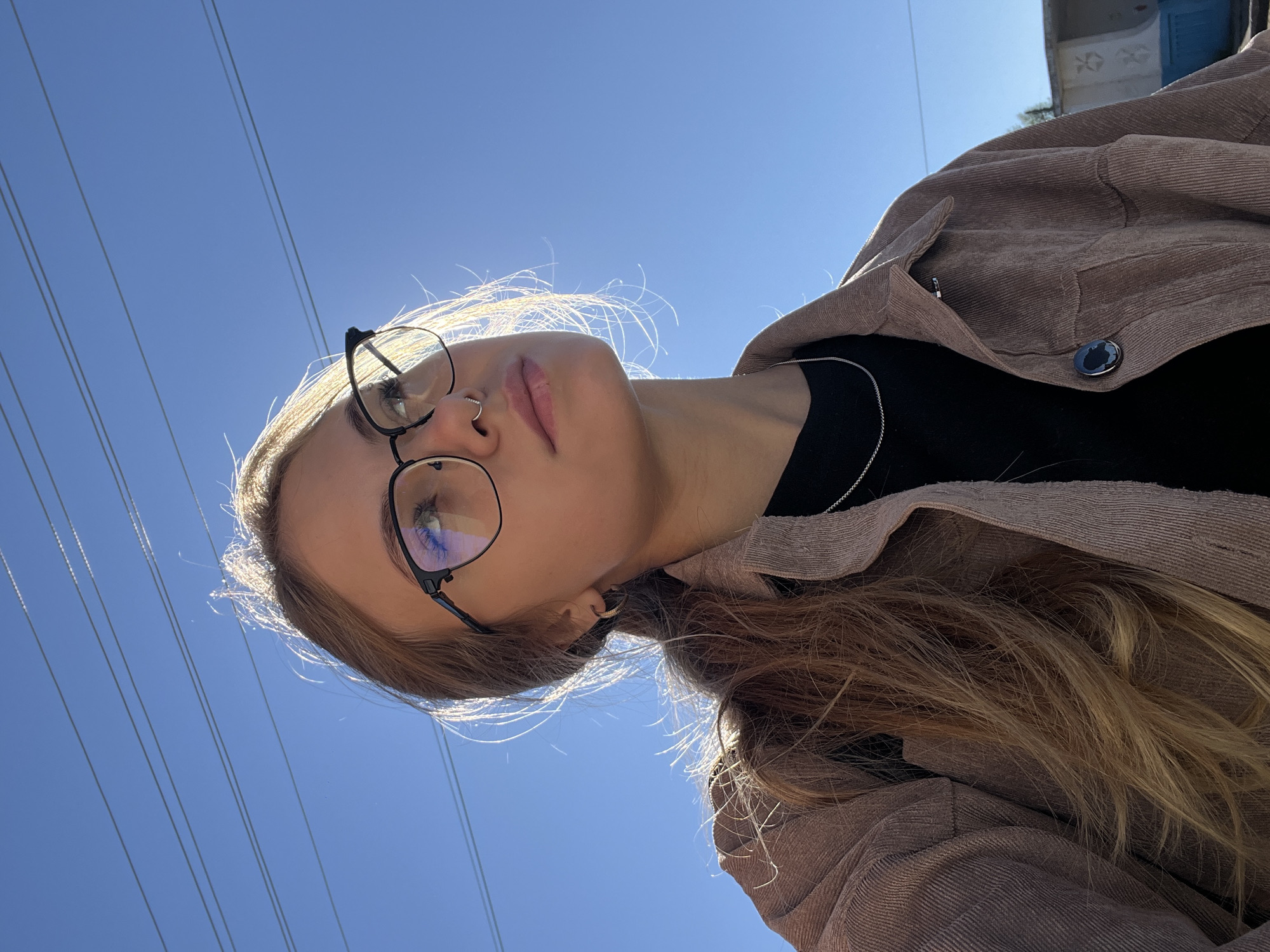 A young woman with blonde hair pulled back in a ponytail, wearing glasses, a nose ring, and a tan corduroy jacket over a black shirt, is looking upwards against a clear blue sky with power lines visible in the background. The lighting suggests it is daytime, with the sun backlighting the subject. The scene appears to be outdoors, possibly in an urban or semi-urban setting given the power lines and a distant building. There is no information in the image to determine the specific location within the city of Фоки, Russia.