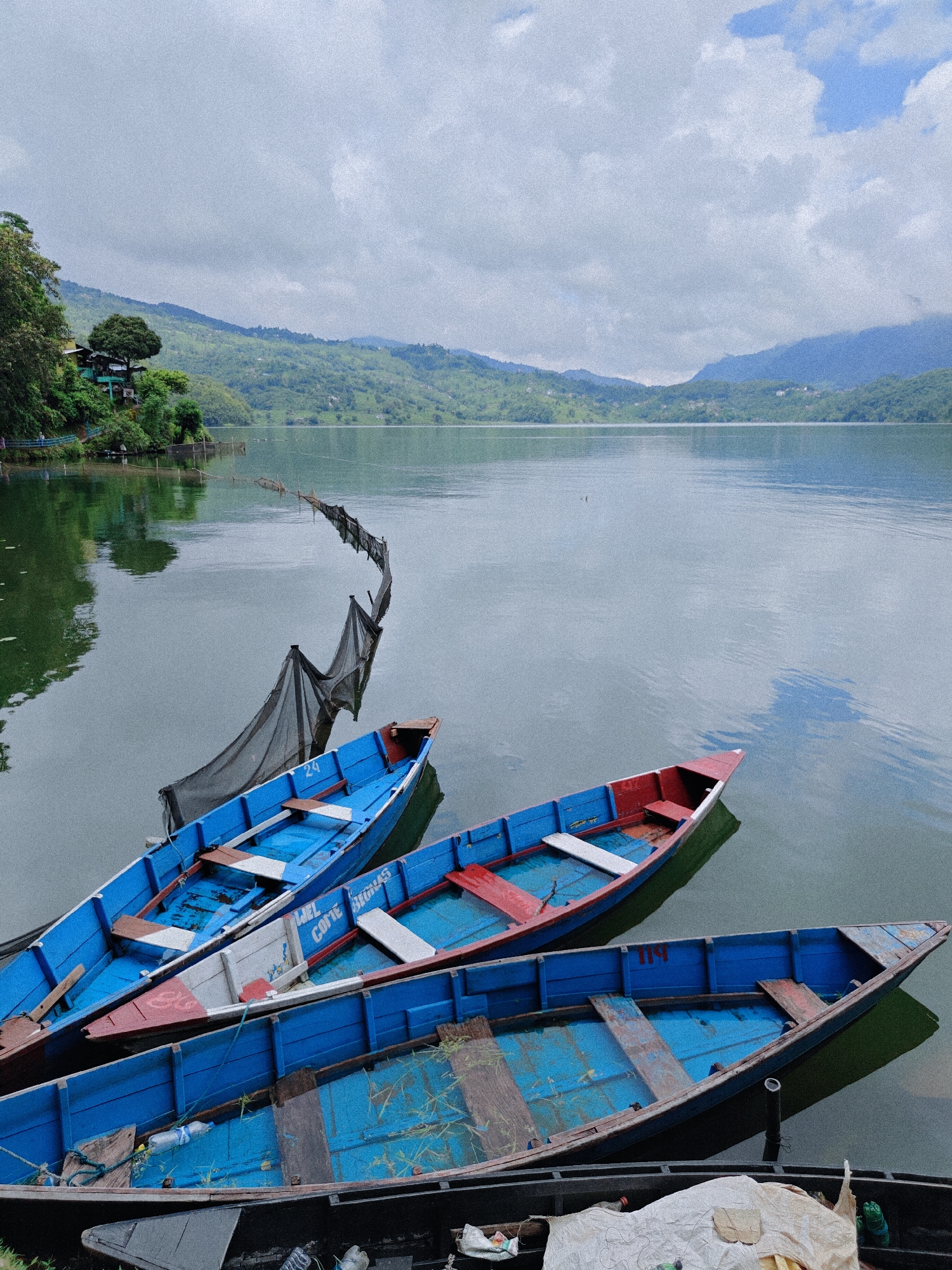 The image depicts a serene lake scene in Pokhara, Nepal, likely Phewa Lake, given the context of the city's geography. In the foreground, several traditional wooden boats are moored, their vibrant blue and red paint contrasting with the calm, muted green and grey waters of the lake. The boats are of varying sizes and states of repair, some appearing more weathered than others. One boat in the middle ground has text on its side that reads "WEL COME BEORRAS", possibly a name or destination.
In the middle distance, a black net, possibly for fishing, is stretched across the water, supported by poles. The net's design is undulating, creating a dynamic element in the otherwise still water.
The background features lush, rolling hills covered in dense greenery, characteristic of Pokhara's landscape. The sky is overcast with a mix of grey clouds and patches of lighter, diffused light, suggesting a soft, possibly humid atmosphere. A few scattered buildings and trees are visible nestled on the hillsides, hinting at human presence and the natural beauty of the region. The overall impression is one of tranquility and natural beauty, with the boats adding a touch of local character to the picturesque setting. There are no people visible in the image.