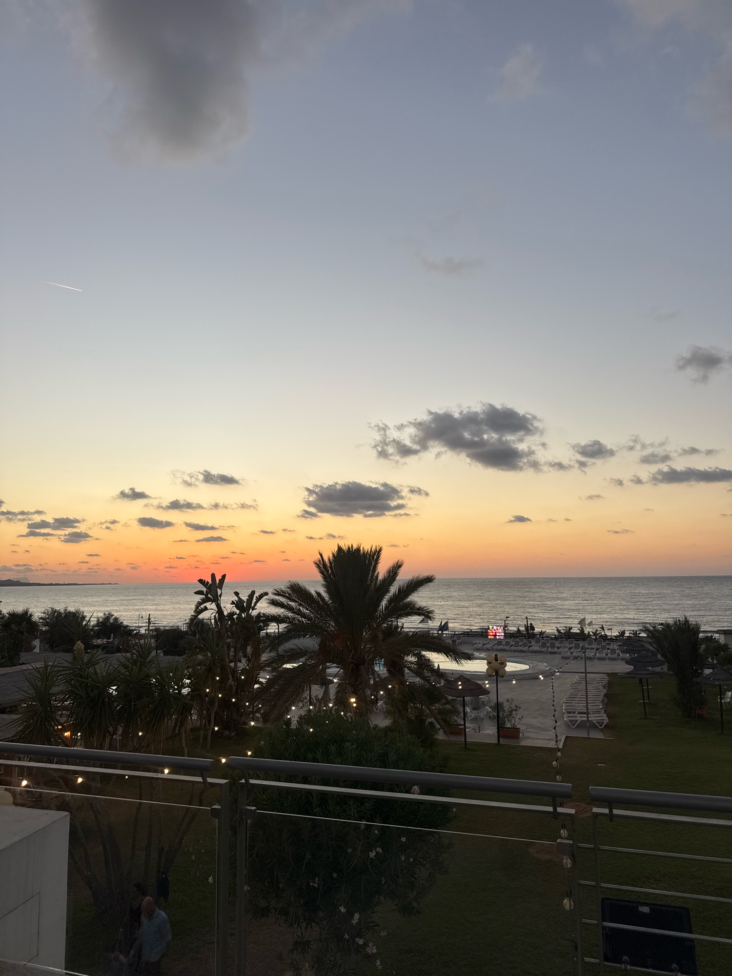 This image depicts a sunset over the sea, viewed from an elevated position, likely a balcony. The sky displays a gradient of colors, from pale yellow at the horizon to a dusky blue higher up, with scattered clouds. A thin, white contrail from an aircraft is visible in the upper left portion of the sky. In the foreground, there is a railing with string lights. Below the railing, a well-maintained lawn is visible, with palm trees and other vegetation. A swimming pool with lounge chairs and umbrellas is situated in the middle ground, adjacent to the beach. In the far distance, the sea meets the coastline, with faint outlines of buildings on land. The overall scene suggests a resort or hotel setting in Isola delle Femmine, Italy, known for its coastal views and sunsets.