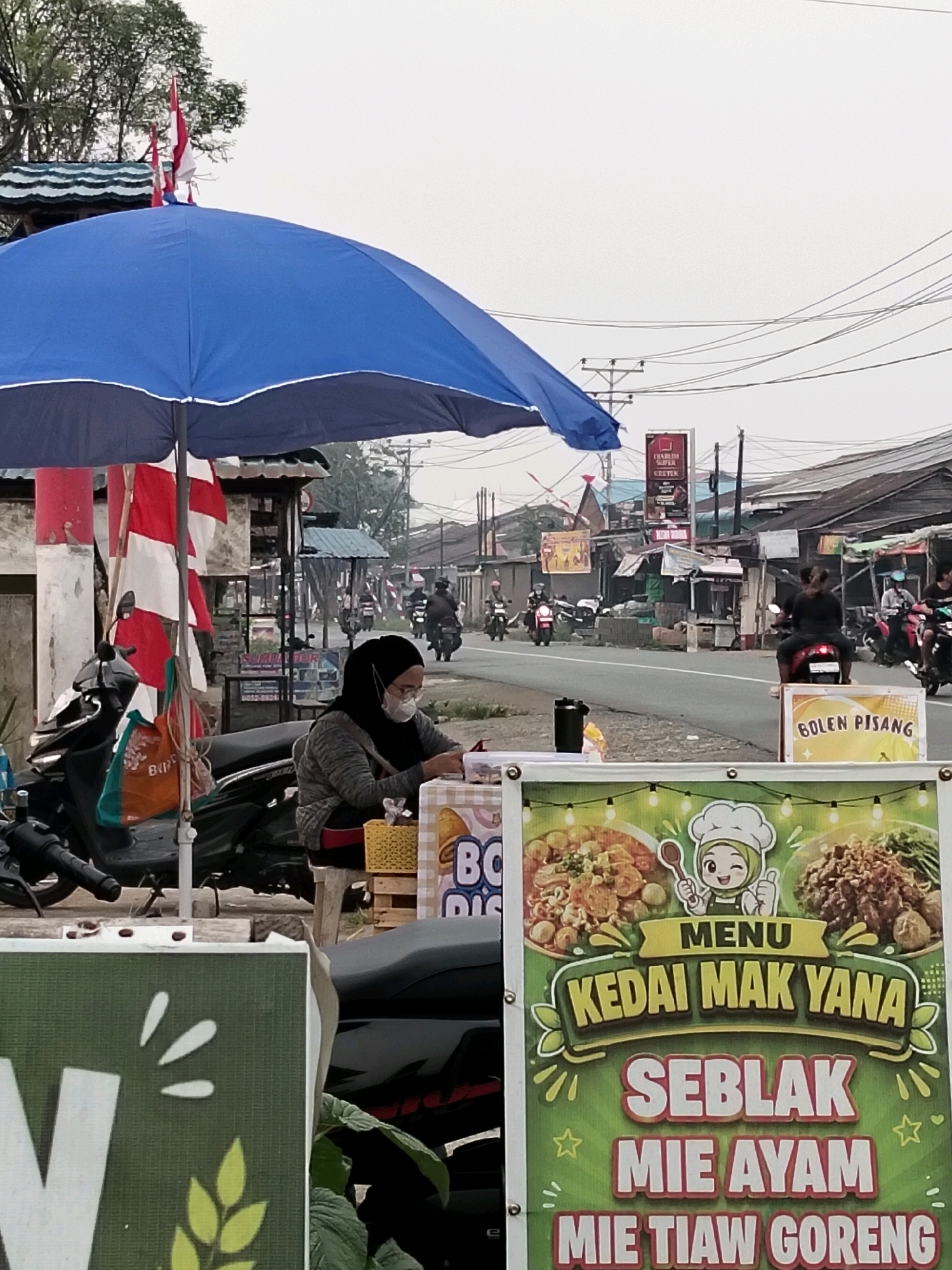 An eye-level, medium shot captures a street-side food stall in Arang Limbung, Indonesia. The stall is situated along a road with several motorcycles visible on the street. The sky is overcast. A large blue umbrella provides shade over the stall. In the foreground, a woman wearing a black hijab and a white face mask is seated behind the stall, seemingly attending to her business. She is wearing a grey sweater and red accents. To her right, a yellow basket sits on top of some wooden pallets. A large green and white sign advertising food items such as "SEBLAK", "MIE AYAM", and "MIE TIAW GORENG" is prominently displayed. In front of the stall, a portion of another green sign is visible with white leaf-like illustrations. A motorcycle is parked to the left of the woman. The background depicts a street lined with buildings and utility poles with numerous wires. Several motorcycles are in motion on the road. A smaller red and white flag is attached to the top of the umbrella.