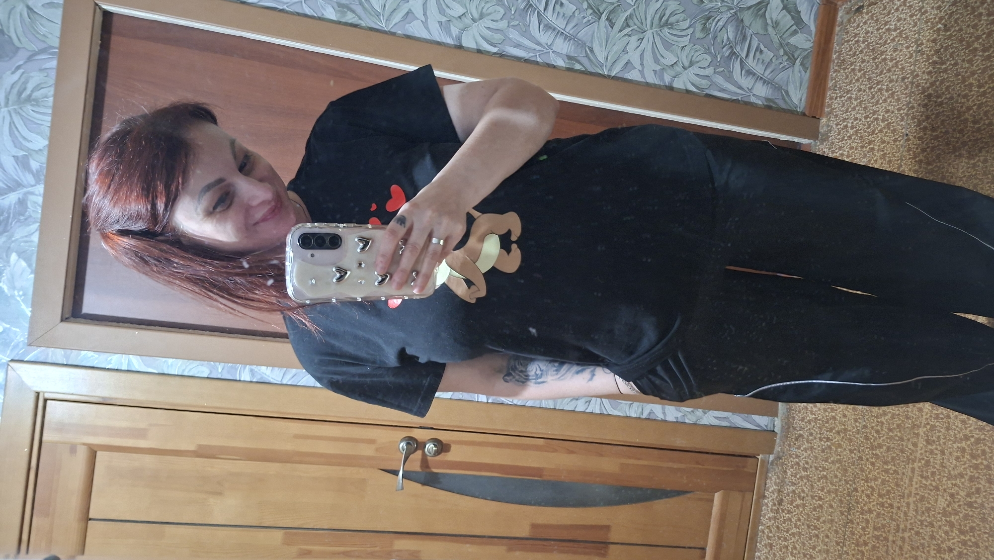 A woman takes a mirror selfie inside an apartment. The woman has red hair and is wearing a black T-shirt with a bear design. She is holding a phone with a clear case decorated with hearts. The location is possibly in Забалоцкі сельскі Савет, Belarus. The background includes a wooden door, a patterned wall, and a glimpse of the floor.