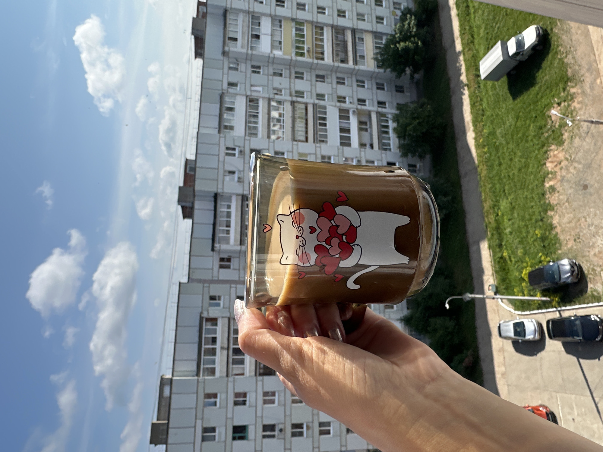 A person's hand with manicured nails holds a clear glass mug filled with a brown liquid, likely coffee, with a white cat graphic holding red hearts on its surface. The mug is positioned in front of a tall, grey apartment building with numerous windows and balconies. In the background, there is a clear blue sky with scattered white clouds. Below the building, a grassy area is visible with several parked cars, including a silver sedan, a black sedan, and a dark-colored SUV. A white truck with a covered bed is parked on the grass. A street light is also visible in the foreground. The scene appears to be an urban residential area in the city of Chaikovsky, Russia, as indicated by the architectural style of the building and the presence of common Russian vehicle models.