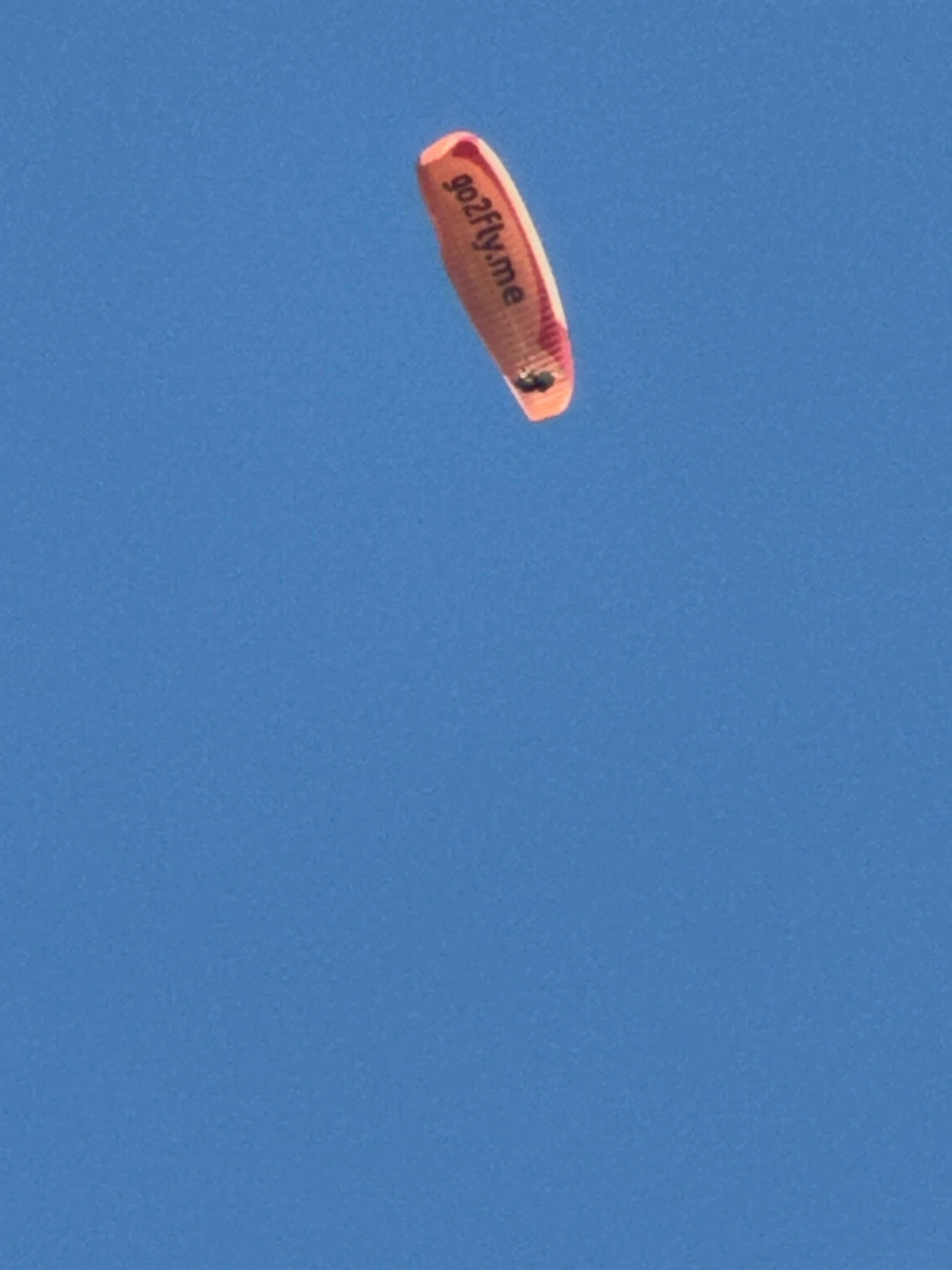 An orange and pink paraglider with "go2fly.me" written in black text is suspended against a clear blue sky. The paraglider appears to be in mid-flight, with dark lines indicating its suspension lines and two black objects that could be harness attachment points. The background is a uniform expanse of blue sky with no discernible geographical features, urban elements, or weather phenomena. The image does not contain information to determine its location in Boreti, Montenegro or any other specific urban setting.