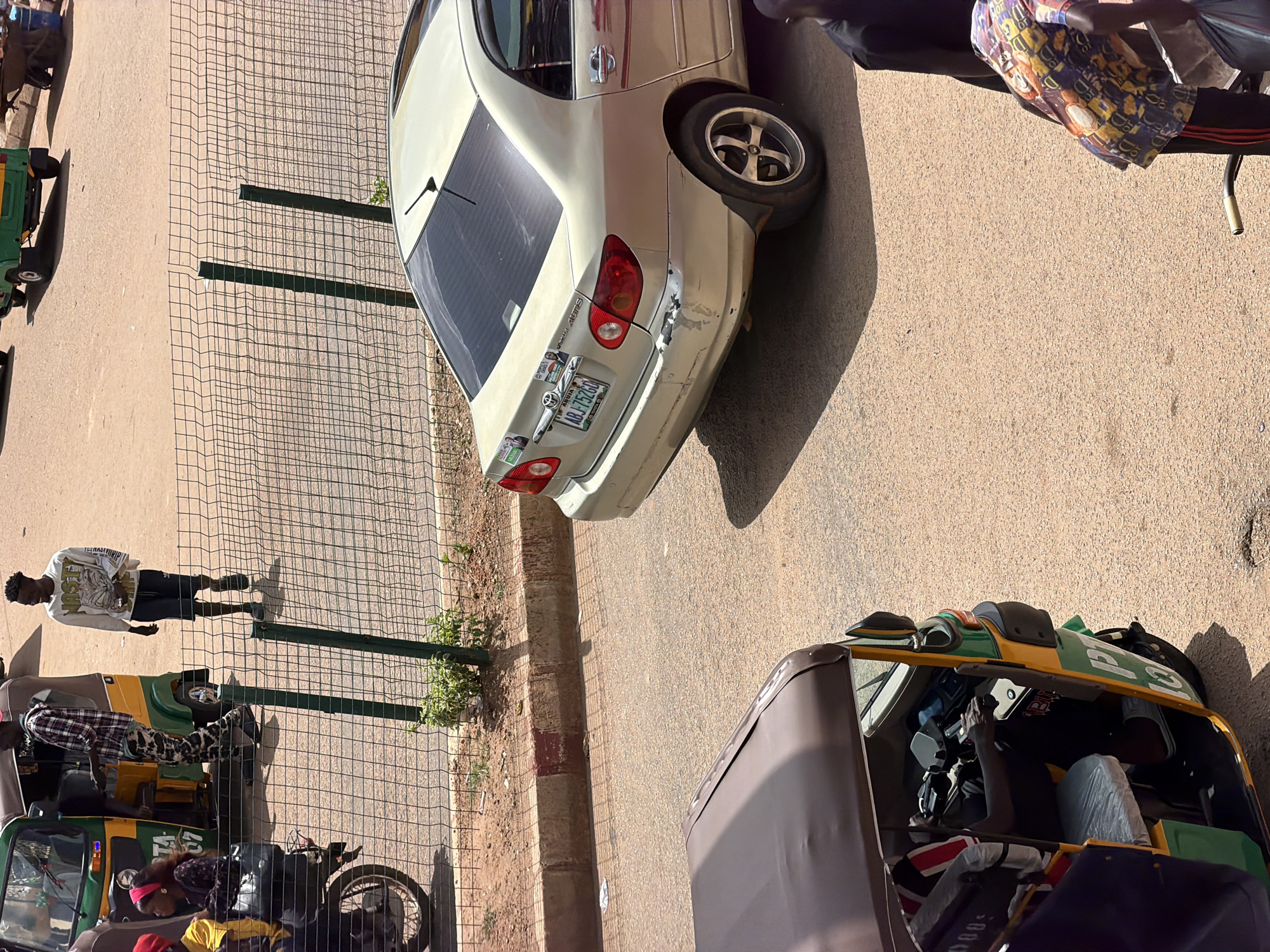 This media file captures an outdoor scene in Bukuru, Nigeria, likely during the daytime given the strong shadows. The image features several subjects and objects arranged on a paved surface with a sandy shoulder and a metal fence.
In the foreground, a brown and green auto-rickshaw (tuk-tuk) is parked. Inside the driver's area, a person is visible, and the interior contains seating with covers and a steering wheel.
Behind the auto-rickshaw, a person stands near a metal fence. This individual is wearing a white long-sleeved shirt with a graphic design and dark pants. Further along the fence line, another auto-rickshaw, this one primarily green and yellow, is parked. A person in a plaid shirt is seated in this vehicle.
Dominating the mid-ground is a light-colored sedan, possibly a Toyota Corolla, parked facing away from the viewer. It displays a license plate that reads "AB 752 GJQ". The car shows some damage on its rear bumper.
The scene appears to be a street or a roadside area, typical of a commercial or residential zone in Nigeria. The weather seems sunny, casting distinct shadows from the vehicles, fence, and people. There is no clear indication of a specific event, but the presence of multiple auto-rickshaws suggests it could be a transport hub or a busy street. The overall impression is one of everyday activity in a Nigerian urban or semi-urban environment.