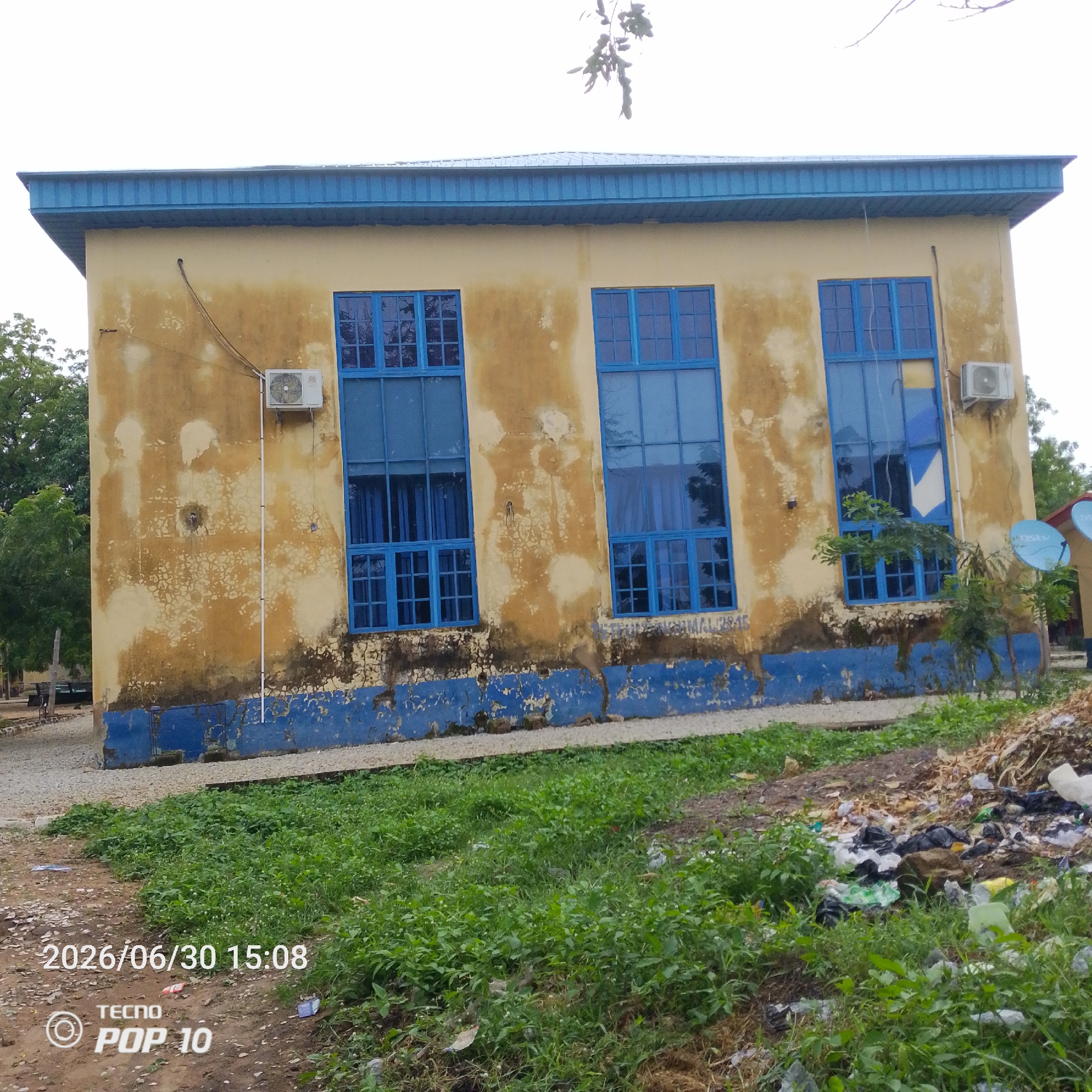 The image shows a building with a faded yellow facade and a blue metal roof. There are three large blue-framed windows on the facade, each divided into multiple panes. An air conditioning unit is mounted on the wall to the left of the rightmost window. The base of the building is painted blue. In front of the building, there is a patch of green grass and some scattered debris, including plastic bags and other waste. The ground in front of the grass is covered with gravel. The timestamp "2026/06/30 15:08" and the text "TECNO POP 10" are overlaid on the bottom left of the image. The scene appears to be in Jalingo, Nigeria, based on the timestamp and inferred location from the context.