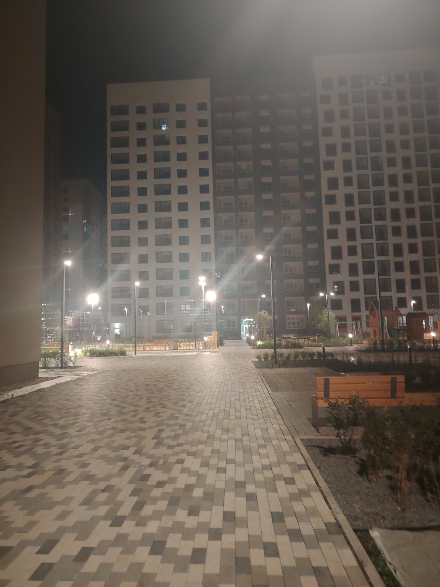 The image depicts a nighttime scene in an urban courtyard. Tall, modern residential buildings with numerous windows dominate the background. The foreground features a paved walkway made of light and dark gray rectangular pavers, arranged in a patterned design. Several tall, modern streetlights illuminate the area, casting a bright glow. Greenery, including bushes and small trees, is visible along the sides of the walkway and around the base of the buildings. In the mid-ground, there are wooden benches, and a children's playground with structures resembling a slide and climbing frame can be seen. The overall atmosphere is one of an illuminated, modern residential complex at night. The image does not contain any individuals or specific actions.