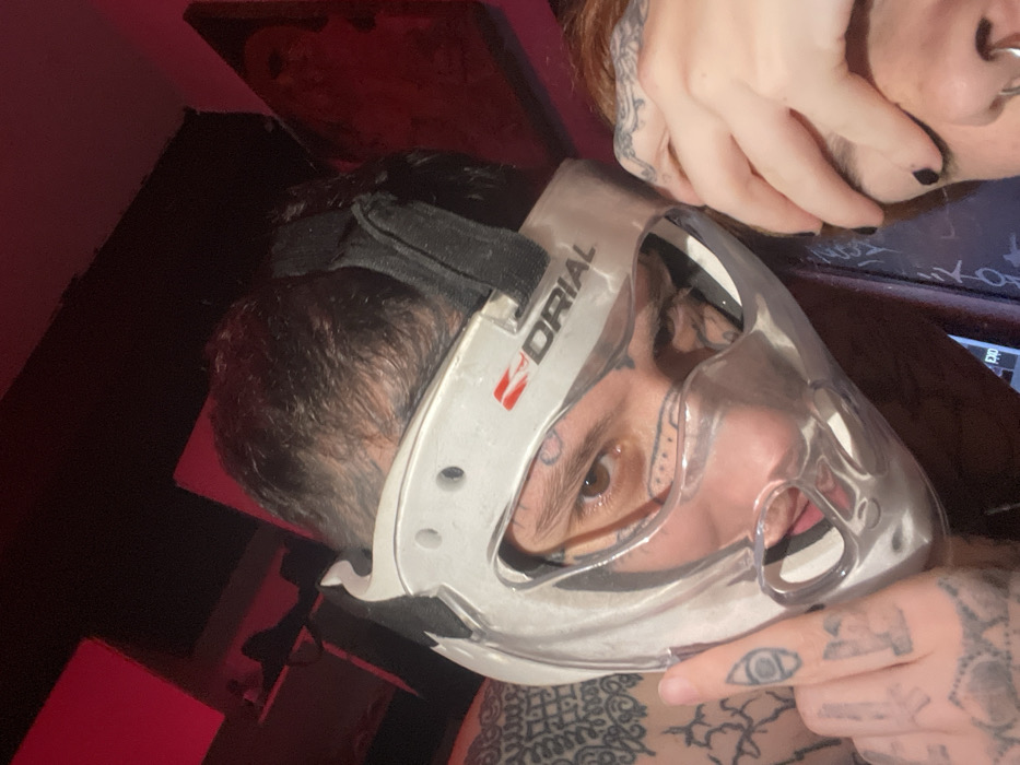 A close-up, slightly distorted image shows a person wearing a clear hockey mask. Visible tattoos adorn the person's face, neck, and hands, including an eye on their index finger. Another individual's hand is present, with black nail polish and tattoos. The background includes a dimly lit room with red lighting, and a framed picture is mounted on the wall. The hockey mask, branded "DRIAL," dominates the view.