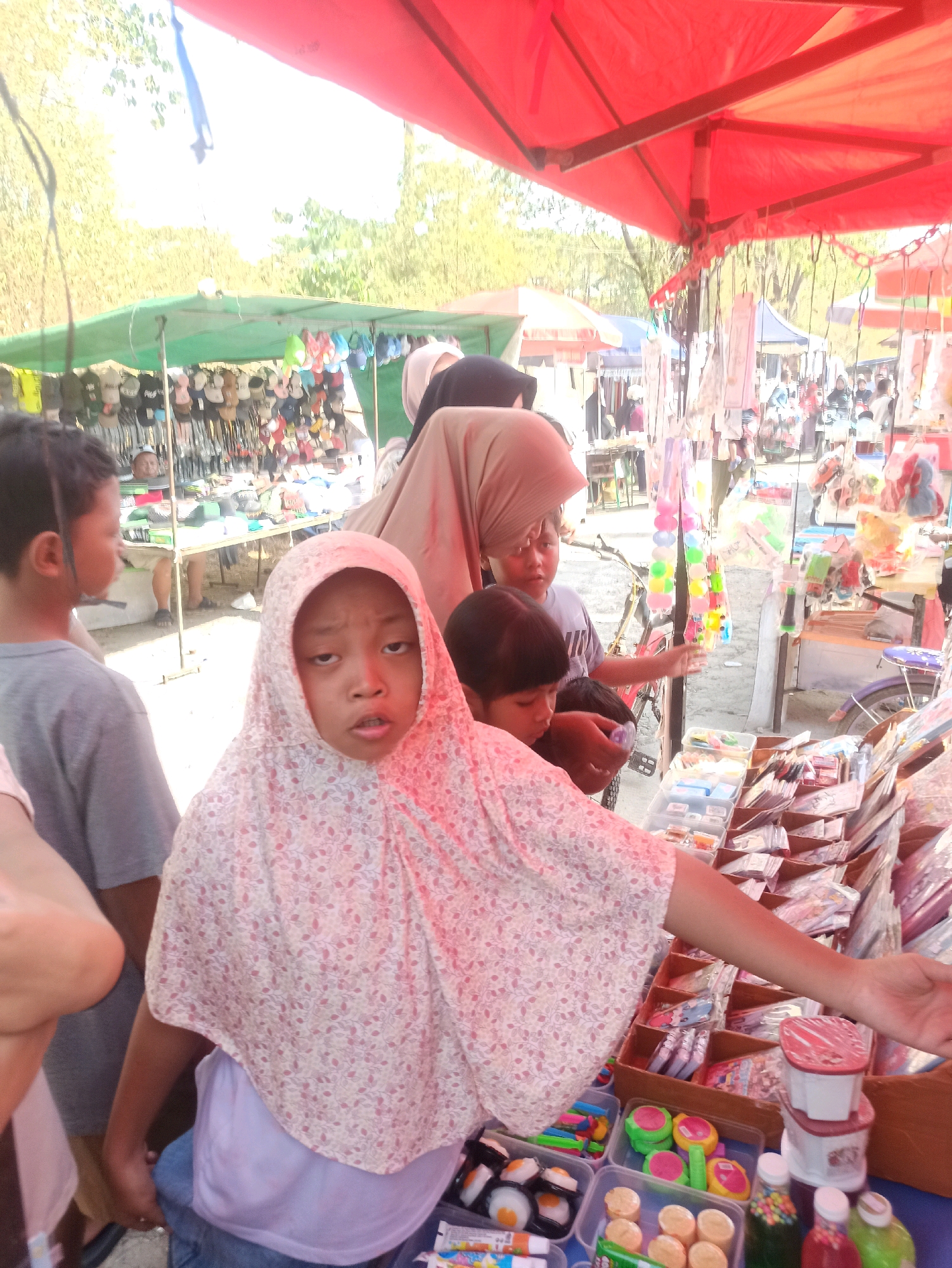 The image depicts an outdoor market scene in Telagamurni, Indonesia, likely a street market or flea market given the temporary stalls and arrangement of goods. The foreground features several children. A young girl wearing a pink floral hijab is prominently positioned, looking towards the viewer with an open mouth. To her left, a boy in a gray t-shirt faces away, observing the market. Another child, partially visible, appears to be interacting with items on a stall.
The stalls are laden with various merchandise. One stall displays an array of items, including what appear to be toys, such as colorful plastic beads and small figurines, as well as packaged goods like stationery and small containers. Another stall, visible in the background, is covered with a green tarp and displays a variety of clothing items, including shirts and hats. The market is covered by red and green tarps, suggesting a temporary setup.
The environment appears to be sunny, with trees and other market stalls visible in the background, indicating a bustling and active commercial area. The overall impression is one of a lively community market.