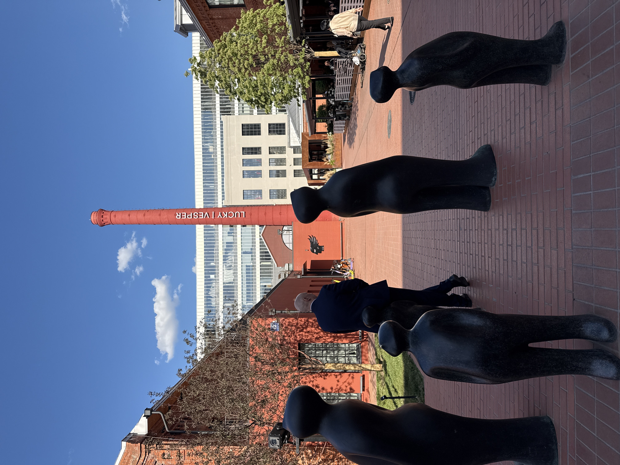 This media file depicts a vibrant outdoor scene in an urban pedestrian area, likely in Moscow, Russia, under clear blue skies. The image features a mix of artistic sculptures, people, historical brick architecture, and modern buildings.
In the foreground and middle ground, several dark, abstract, anthropomorphic statues are arranged along a pathway paved with reddish-brown bricks. These statues are elongated and featureless, casting long shadows. A man in a dark suit is seen walking past one of these statues. Further back, other individuals are visible, including one person in a light-colored jacket walking, and several others seated at outdoor dining areas with white fencing.
The background is characterized by a blend of architectural styles. A tall, red brick industrial chimney stands prominently, bearing the white vertical text "LUCKY | VESPER." Surrounding it are other red brick buildings with various windows, some arched. Behind these, a large, modern high-rise building with a glass facade reflects the bright sky. A stylized black bird logo is painted on a red wall in the distance. Sparse trees suggest either late autumn or early spring.
The time of day is clearly daytime, with bright sunlight and a few scattered white clouds in the sky, indicating fair weather. The overall impression is one of an active public space combining industrial heritage with contemporary urban life and art.