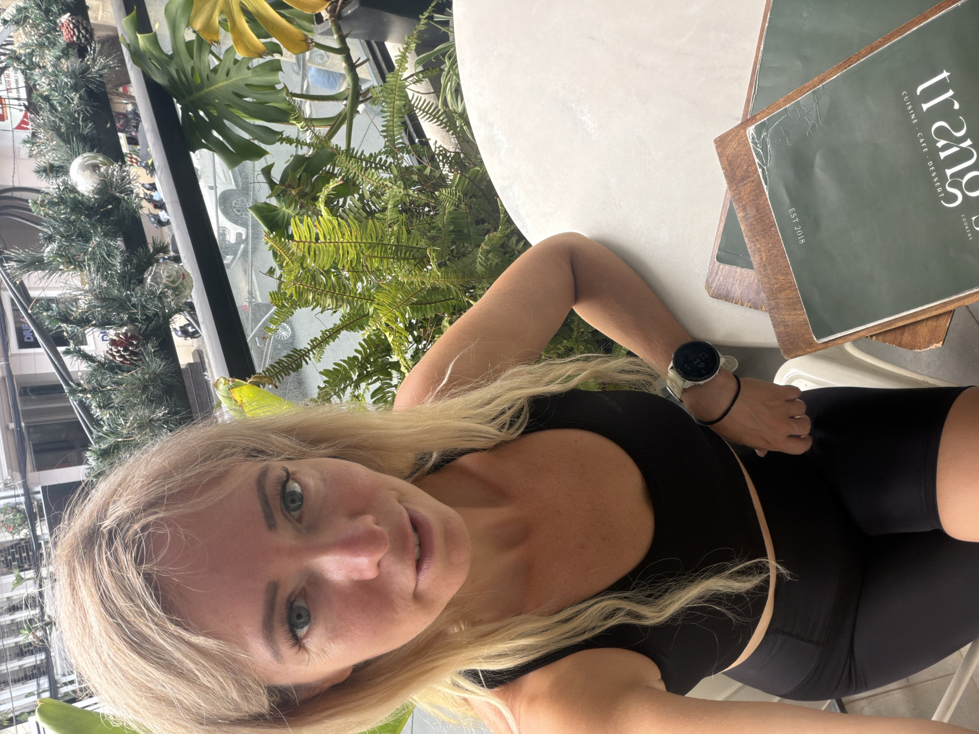 A blonde woman with blue eyes and fair skin is taking a selfie in an outdoor setting, possibly a cafe or restaurant balcony in Da Lat, Vietnam. She is wearing a black sports bra and black shorts. She has a smartwatch on her left wrist. In front of her, on a white table, are two menus. One menu is green with "trang" in white lettering and "CUISINE - CAFE - DESSERT" and "COOKERY" in smaller white lettering. It also has "EST. 2018" printed on it. Behind her, there are plants and a railing overlooking a street with parked motorbikes and a white car. Christmas decorations, including pinecones and silver ornaments, are visible on the railing. The lighting suggests it is daytime.