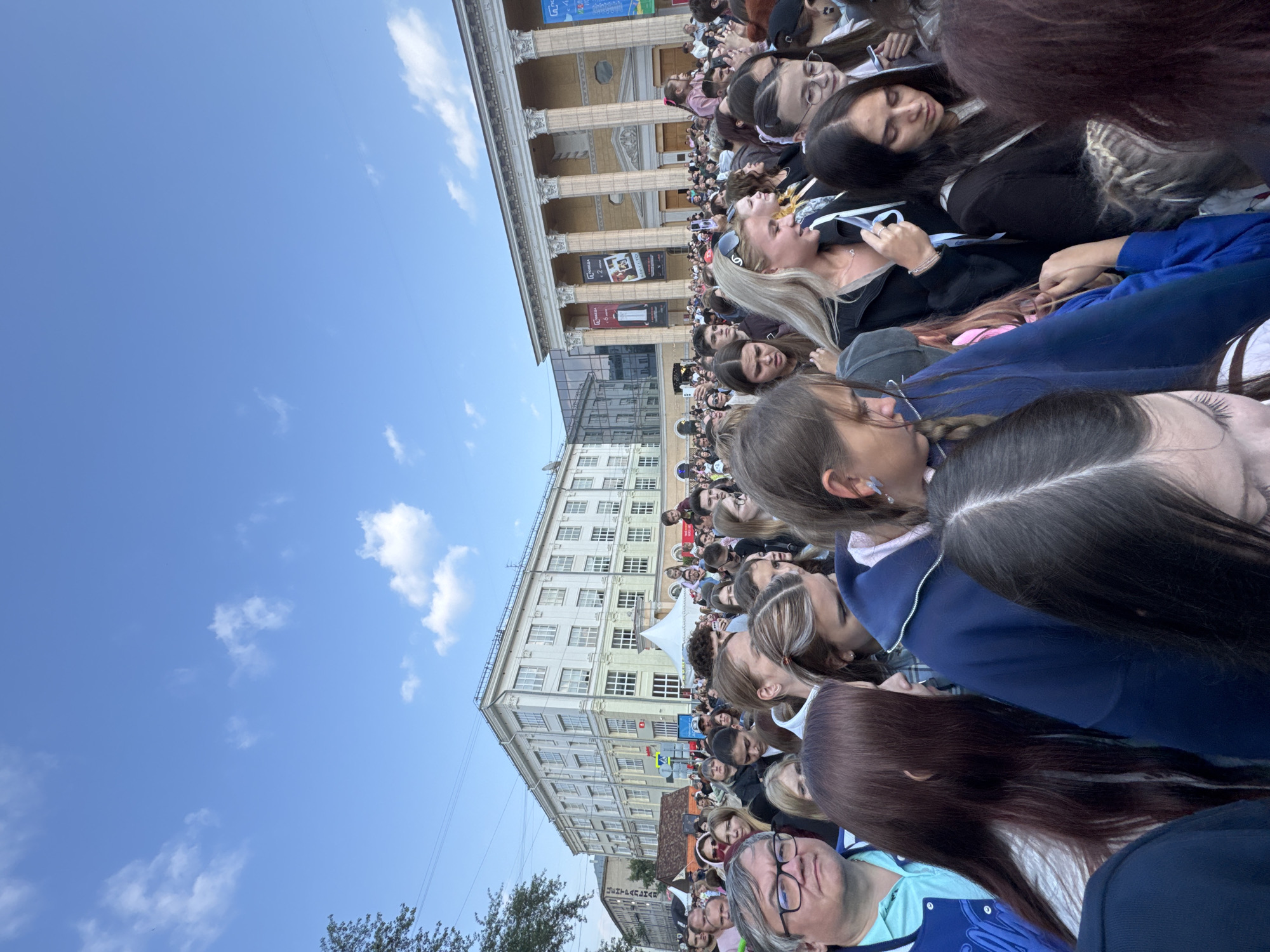 A large crowd of people, predominantly young adults, are gathered outdoors in a city square. The sky above is blue with scattered white clouds. In the background, there are two prominent buildings. The building on the left appears to be a multi-story, light-colored structure with numerous windows. The building on the right is larger and features a classical facade with a colonnade. Banners are visible on the right building, some of which appear to be advertising events. Several white tents are set up in the middle ground. The people in the foreground are tightly packed together, suggesting a concert, festival, or public gathering. They are facing in various directions, with many looking forward, presumably towards a central stage or event area. Some individuals are holding up their phones, possibly to record or take pictures. The overall atmosphere seems lively and engaged. The location is the Lenin Square in Novosibirsk, Russia.