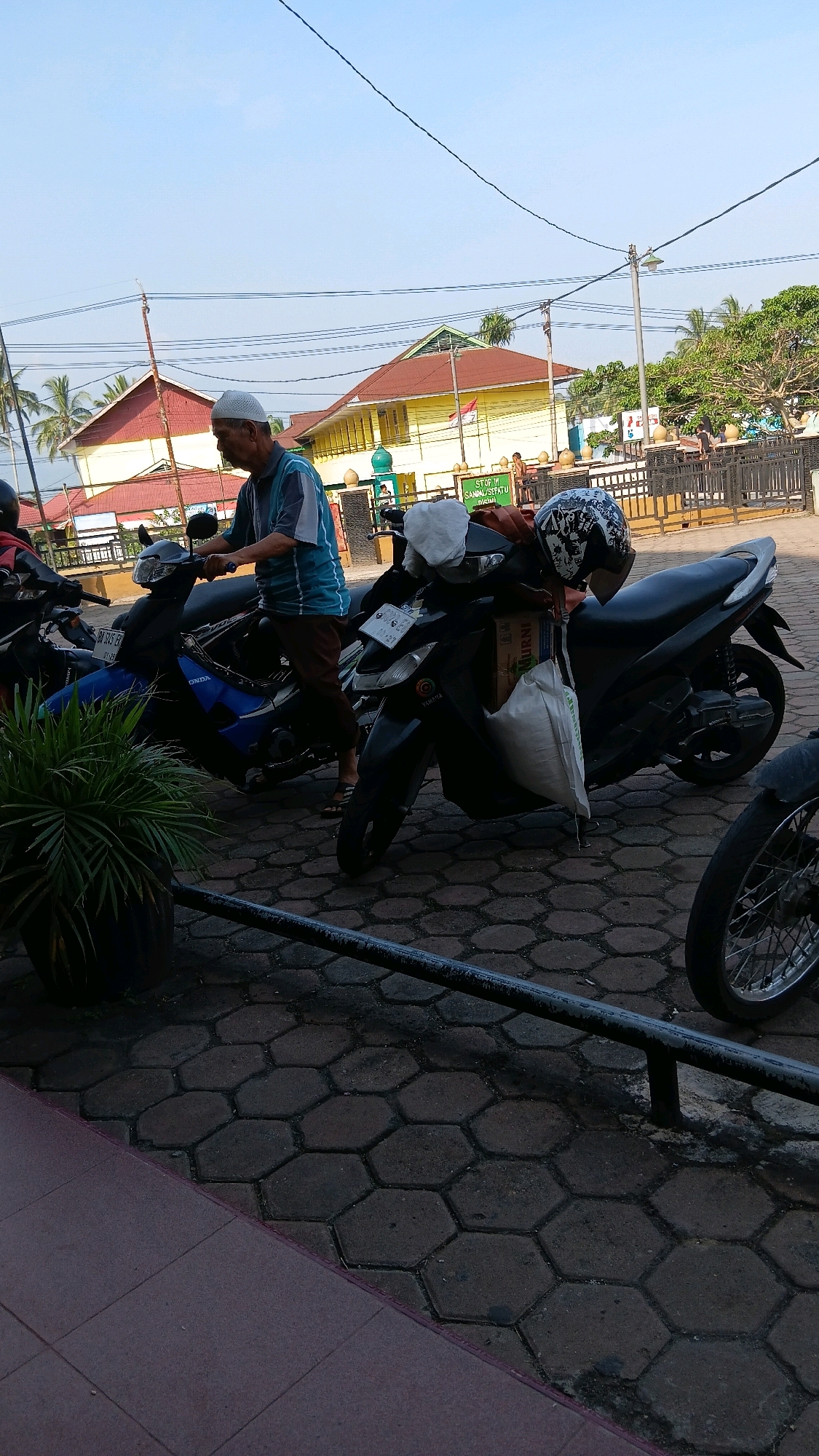 In Sungai Tarab, Indonesia, an elderly man wearing a white cap and a blue and white striped shirt is seated on a dark-colored scooter. He is looking towards the right side of the frame. Beside him, another scooter is parked, which has a white bag and a helmet with a black and white pattern attached to it. In the background, there are buildings with yellow and red roofs, palm trees, and a clear blue sky. A black metal railing is visible in the foreground, and the ground is paved with hexagonal bricks.