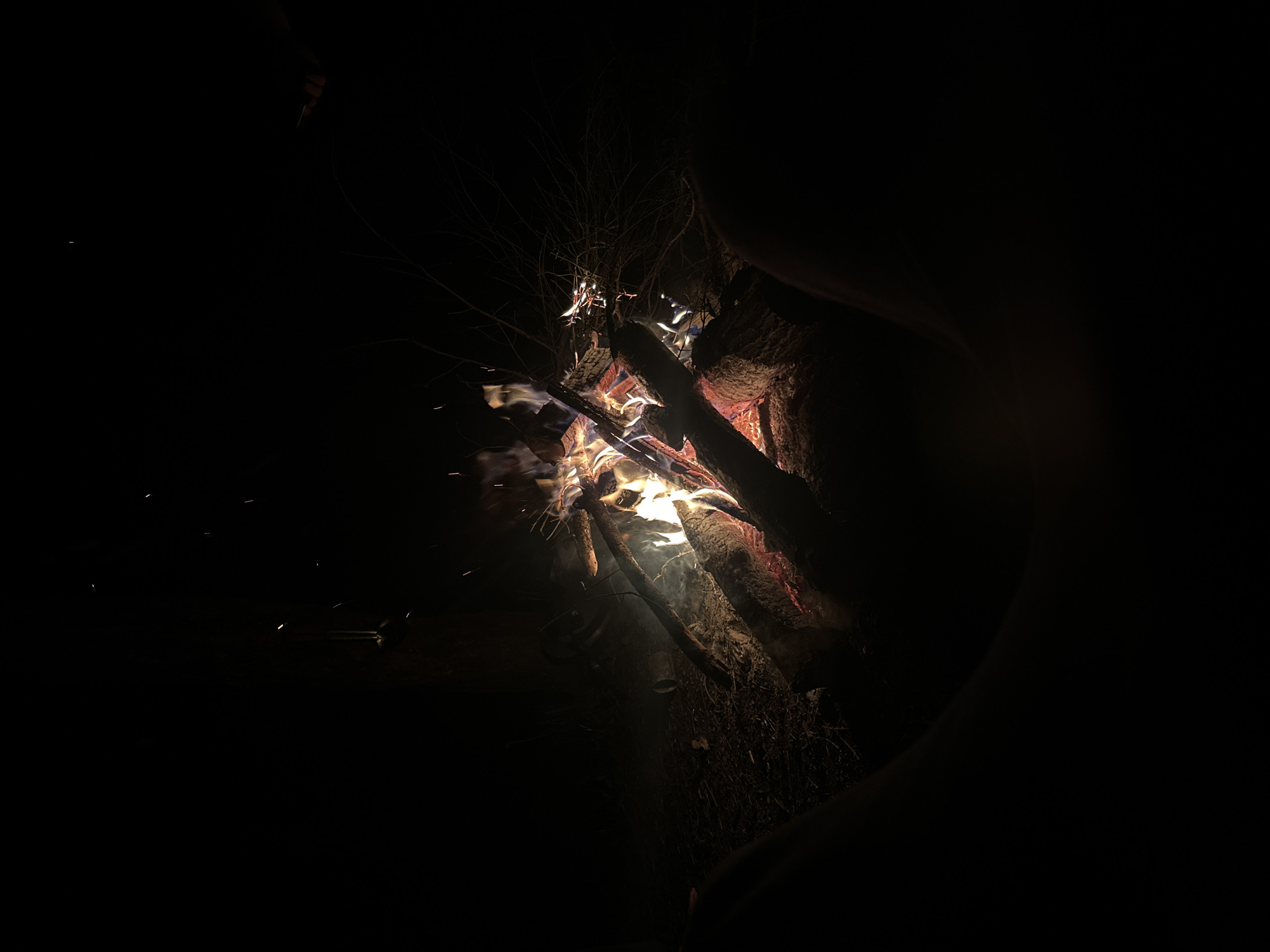 The image depicts a small bonfire in a very dark environment. Logs and twigs are visible within the fire, some appearing to be actively burning with flames and sparks. The background is black and indistinct, suggesting nighttime. The location of Юзуфоўскі сельскі Савет, Belarus cannot be determined from the image alone.