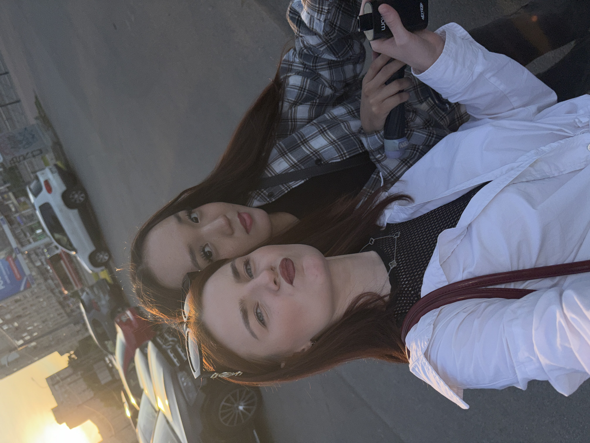 Two young women are taking a selfie outdoors, with a city street in the background. The woman in the foreground has auburn hair and is wearing a white shirt over a black mesh top, with a dark red purse strap visible. She is making a pouty face and has sunglasses perched on her head. The woman in the background has long dark hair and is wearing a plaid shirt over a black top. She is holding a black cylindrical object. Cars are parked along the street, and buildings are visible in the distance, suggesting an urban environment. The sun is setting, casting a warm glow on the scene. The location is the city of Sinyavino, Russia.