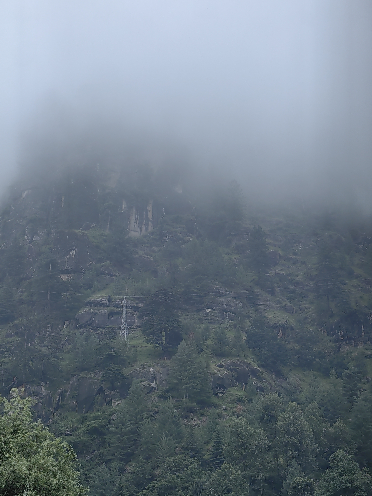 The image shows a mountain shrouded in thick fog. The lower part of the mountain is covered with dense green trees, while the upper part is rocky with sparse vegetation. A tall, metal electrical pylon is visible on the mountainside, indicating the presence of infrastructure. The sky is completely obscured by fog, giving the scene an ethereal and mysterious atmosphere. The location is identified as Bahang, India.