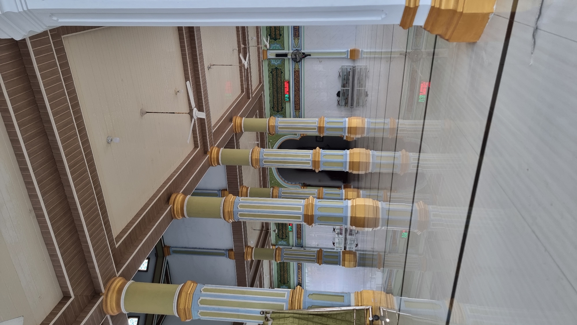 The image depicts the interior of a mosque, characterized by a series of tall, decorative columns supporting an ornate ceiling. The columns are painted with a repeating pattern of yellow, blue, and light green, topped with golden capitals. The ceiling features a combination of wooden and cream-colored panels, with several ceiling fans installed. The floor is highly reflective, likely polished marble or tile, mirroring the columns and ceiling. In the background, there are arched doorways and a digital display showing the time "17:20" in red digits. A white cabinet with glass doors is visible, along with a black standing fan. The overall impression is one of spaciousness and intricate architectural design, consistent with a place of worship. The location is identified as Meunasahlhok, Indonesia.
