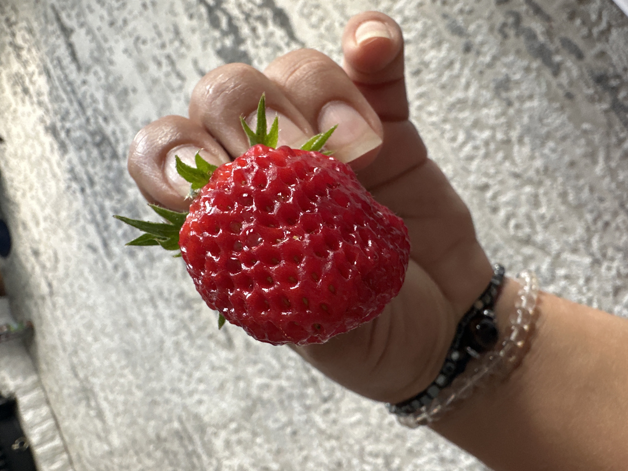 A close-up, slightly overhead shot captures a person's hand holding a single, bright red strawberry. The strawberry, glistening with moisture, is plump and features visible seeds on its surface, with two small green leaves extending from its stem. The hand holding the fruit shows a light-to-medium skin tone, with manicured fingernails that are a pale, translucent color. The person is wearing two thin bracelets on their wrist: one with dark beads and a small, dark pendant, and another with clear, small beads. The background is a textured, light-colored surface, possibly a floor or countertop, with a gray, marble-like pattern. The lighting suggests an indoor setting with natural light.