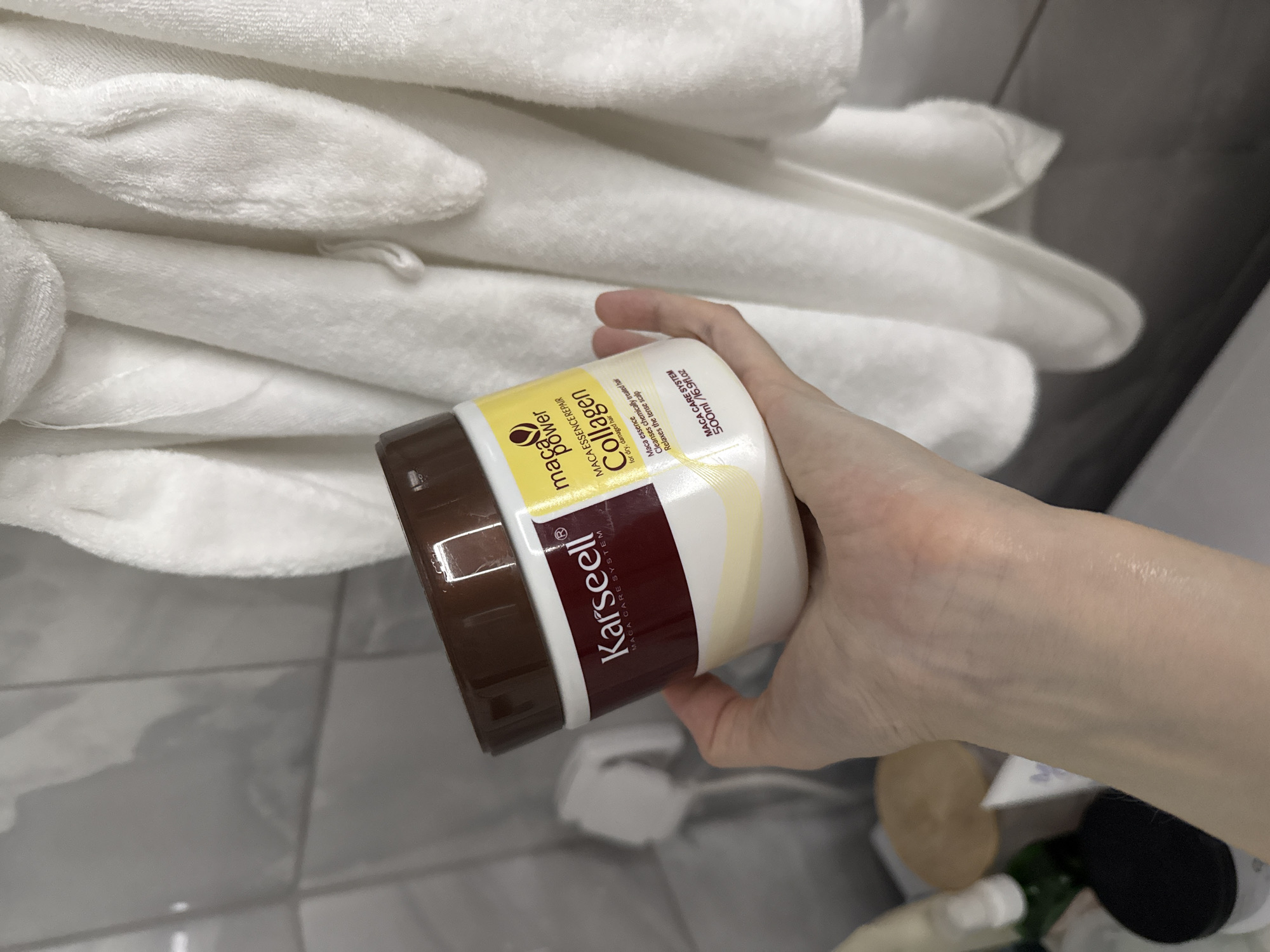 A person's hand is holding a jar of Karseell Maca Power Collagen hair mask in front of a stack of white towels. The jar has a brown lid, a white label with maroon and yellow accents, and text in English describing the product as a hair mask for dry, damaged hair that cleanses chemically treated hair and relaxes the tense scalp. In the background, there is a gray tiled surface and several unidentifiable bottles and containers. The location is not identifiable as Izhevsk, Russia, based solely on the image.