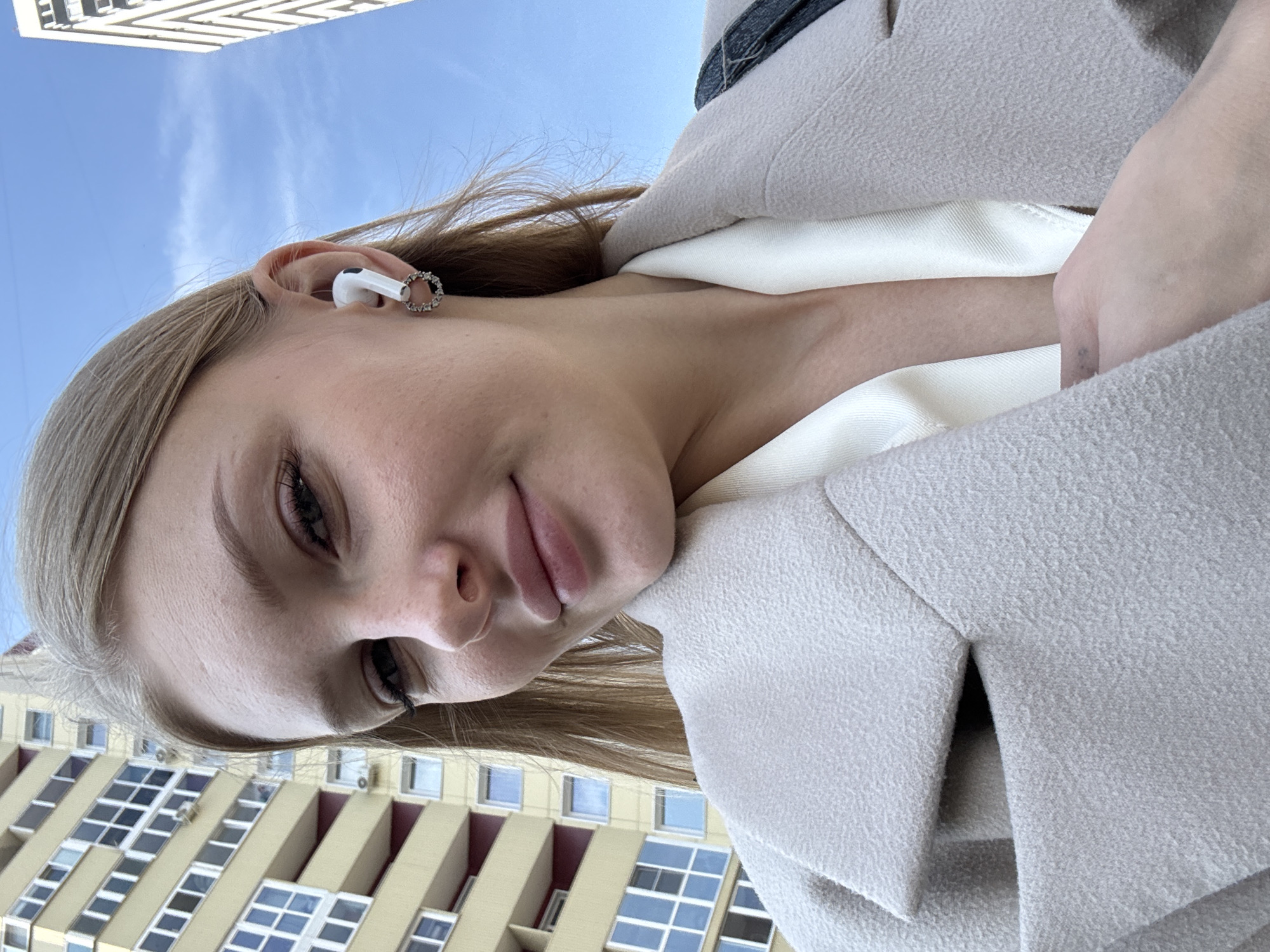 A young woman with blonde hair is taking a selfie outdoors in an urban environment. She is wearing a cream-colored coat and a white top, with a white wireless earbud in her right ear and a circular earring. In the background, a tall, light-yellow apartment building with many windows is visible on the left, and a modern white skyscraper is on the right. The sky is blue with some clouds. The image appears to have been taken during the day. The location is the city of Perm, Russia.