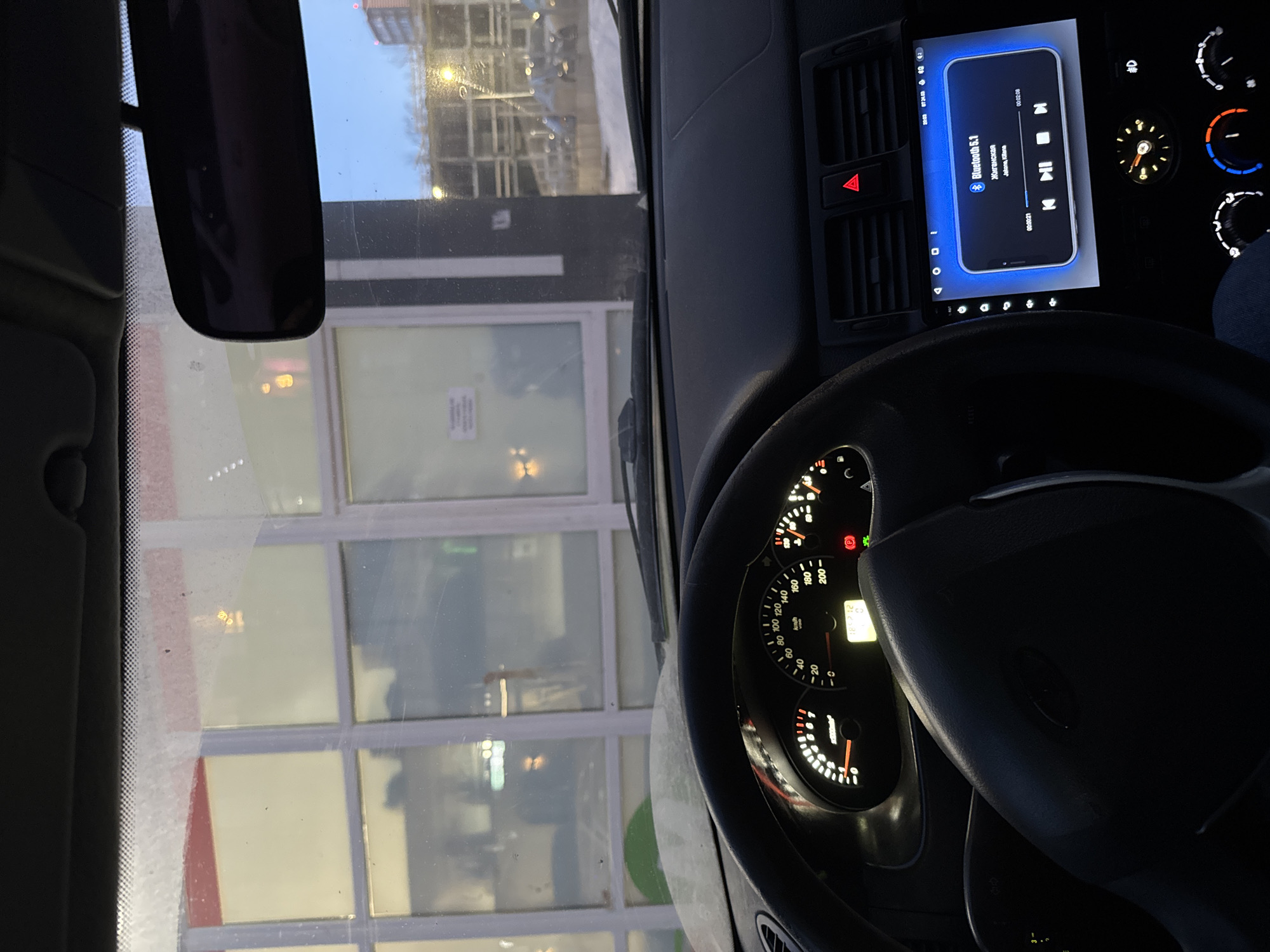 The image depicts the interior perspective from a dark-colored Lada vehicle, likely stationary, looking through its windshield at an urban commercial environment during the evening or night in winter conditions.
The vehicle's dashboard is prominent, featuring an illuminated instrument cluster displaying a speedometer showing 0 km/h, a tachometer, fuel, and temperature gauges. A digital readout indicates "16.12.23" (December 16, 2023) and "0." The steering wheel is dark and partially obscures the lower left of the dashboard. A red triangular hazard light button is illuminated on the center console, alongside air vents. Below these is a modern aftermarket infotainment system, brightly lit, displaying "Bluetooth 5.1" and currently playing the Russian song "Жиганская" by "Jahna, Kitana," with a timestamp of 00:00:21. Below the screen are an analog clock and climate control knobs, one illuminated blue. The rearview mirror is visible at the top center, reflecting dimly lit shapes. The windshield exhibits some dirt or frost.
Outside the vehicle, directly ahead, stands a large commercial building characterized by expansive windows set within white frames, topped by a red horizontal band. Interior lighting from the building is visible through these windows, and a sign with illegible text is partially discernible. To the right of this building, a multi-story parking garage or structure is visible under artificial lighting. The ground throughout the exterior scene is covered in snow, indicating winter. The sky above is a dark blue, typical of dusk or nighttime, with scattered bright streetlights illuminating the area. The scene is located within a commercial district of Perm, Russia, specifically in front of a large retail establishment with an adjacent multi-level parking facility.