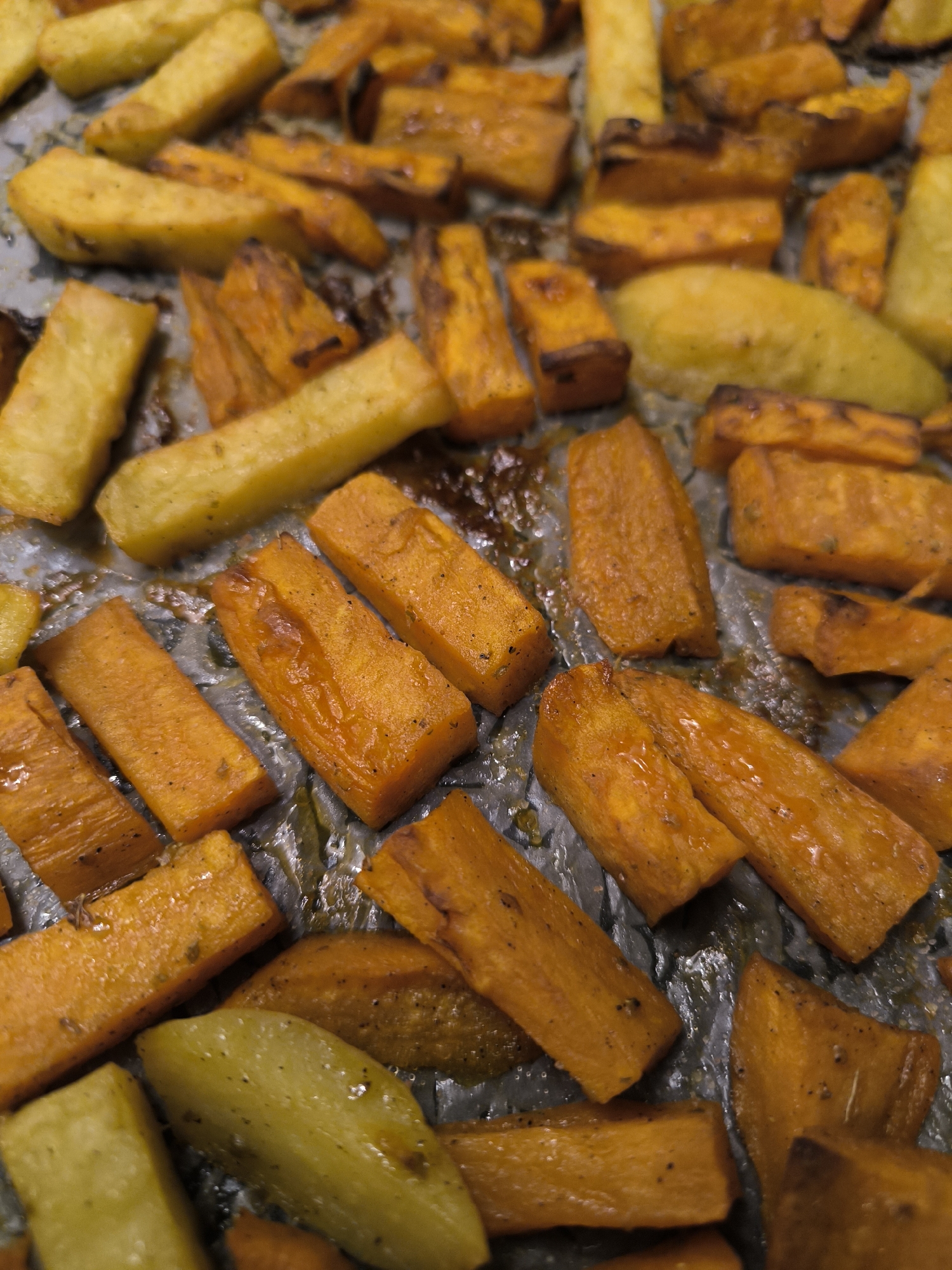 The scene, understood to be situated in the city of Bar, Montenegro, presents a close-up, overhead view of roasted vegetables on a baking sheet. The surface is densely covered with numerous irregularly shaped, elongated pieces of food. Prominently featured are orange-hued segments, consistent with cooked carrots, and lighter yellow/golden segments, indicative of roasted potatoes or parsnips. These vegetables display a semi-glossy sheen from residual oils or moisture, with some edges exhibiting darker browning. Black specks, characteristic of ground pepper or dried herbs, are visible across the surfaces. The metallic baking sheet itself appears dull silver-gray, with dark, viscous residues clinging to areas between the food items. No human presence, explicit actions, or direct interactions are depicted.