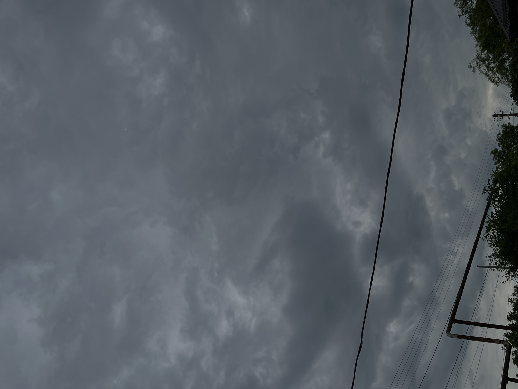 The image depicts an overcast sky with dark, heavy clouds, suggesting impending rain or stormy weather. Two small, bright pink lights are visible in the upper right quadrant of the sky, their source and nature are unclear. In the foreground, thick black electrical wires run horizontally across the frame. Below the wires, a rusty metal pipe structure is partially visible, along with the tops of green trees. The overall scene appears to be an outdoor environment in a residential area, possibly on the outskirts of Novopokrovskaya, Russia, given the context.