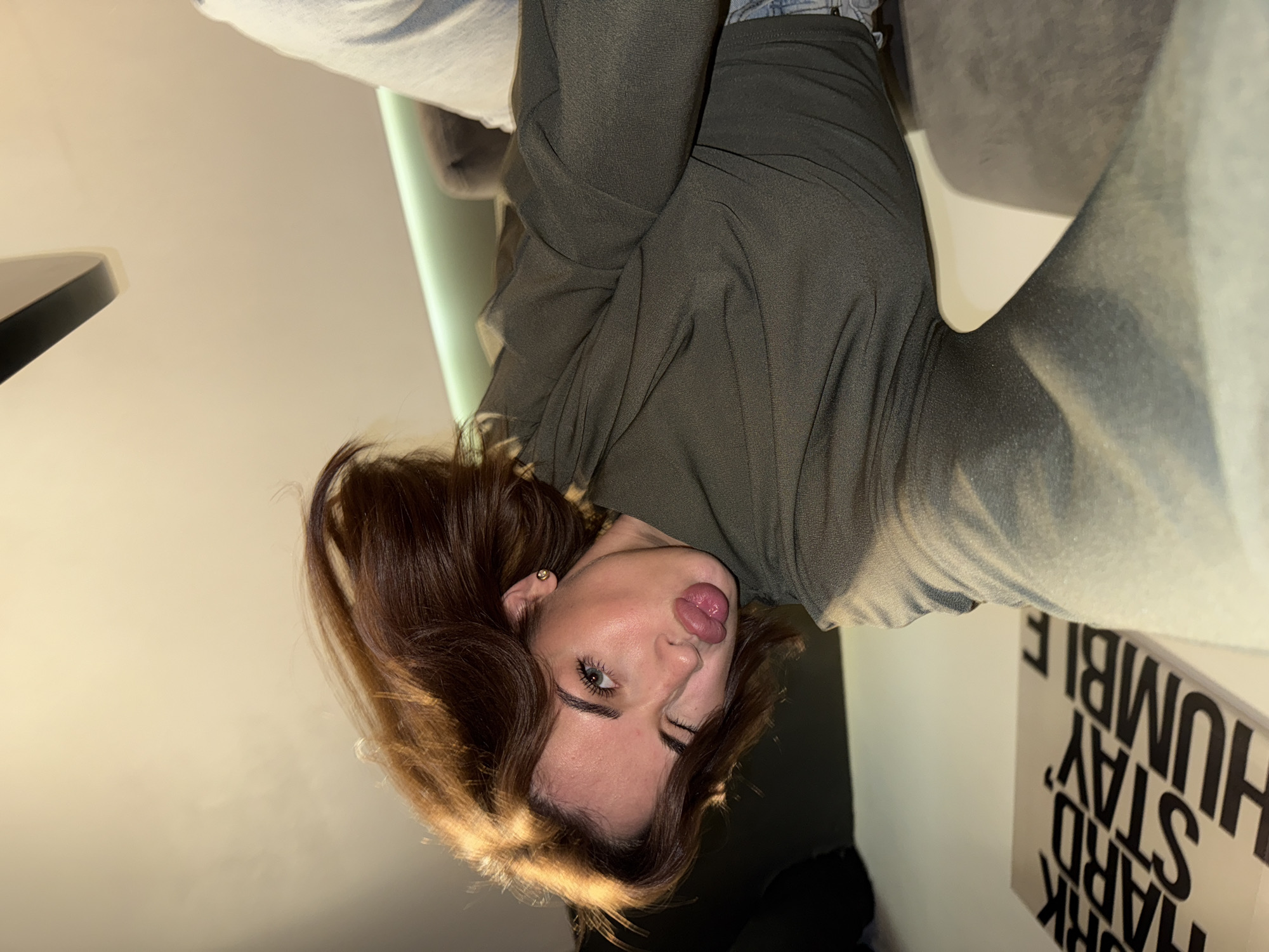 A young woman with brown hair and light eyes is indoors, positioned upside down in the frame. She is wearing an olive-green sweatshirt and light-colored pants. She appears to be making a playful face. The location of the scene is Chaikovsky, Russia. A sign with the words "WORK HARD, STAY HUMBLE" is partially visible. There is a table and a chair in the background. The scene is well-lit.
