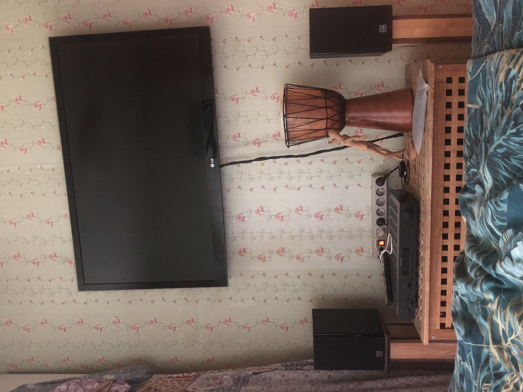 The image depicts a room with floral wallpaper. On the wall, a flat-screen television is mounted. Below the television, on a wooden cabinet, are two black speakers, one on each side. In the center of the cabinet, there is audio equipment, a power strip, a remote control, and a decorative wooden drum with a black net. A small wooden mannequin is positioned near the drum. To the left of the television, a patterned curtain is visible. In the foreground, a bed with blue and green patterned bedding is present. The scene appears to be an indoor residential setting. There is no visible indication of the location being Molgovo, Russia.