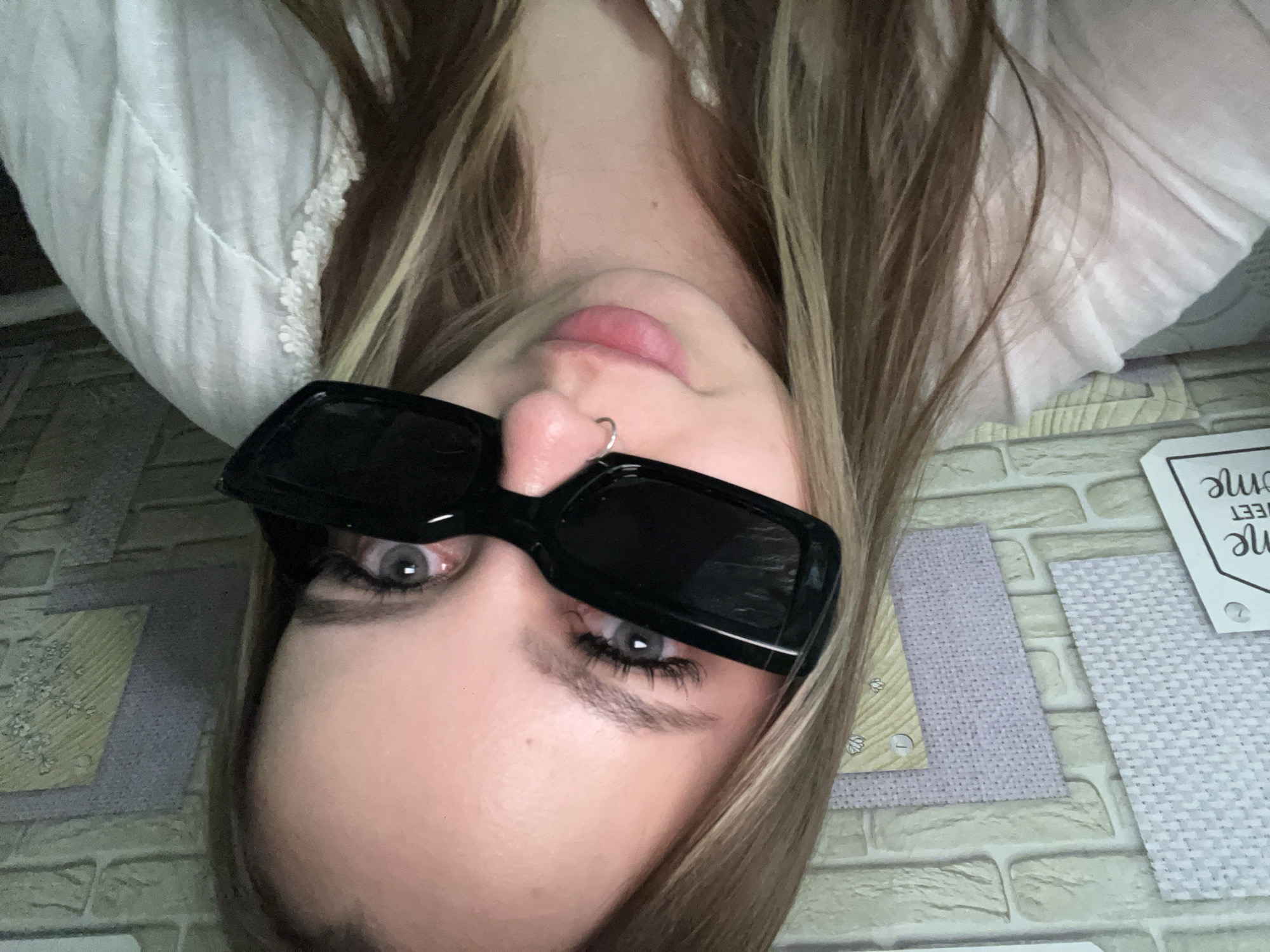 A close-up, inverted portrait features a young person, likely female, with long, light brown and blonde hair framing her face. Her light-colored eyes, possibly grey or blue, are visible beneath black, rectangular sunglasses that are pushed up onto her nose. She wears mascara and has well-defined eyebrows. A small silver hoop nose ring is present on her right nostril, and her pink lips are slightly parted.
She is dressed in a white garment, possibly a shirt or dress, with a delicate lace or embroidered trim visible near her shoulder. The background is a flat surface, patterned to resemble light-colored brickwork, upon which several textured fabric or paper swatches in shades of light purple and light grey are arranged. One swatch shows a small detail with what appears to be the letter 'L' or number '1'. To the far right, a white tag or card is partially visible, bearing the sideways text "SWEET home". The scene suggests an indoor, casual setting, possibly a home or personal workspace in Port-Katon, Russia. The person is looking directly at the camera.