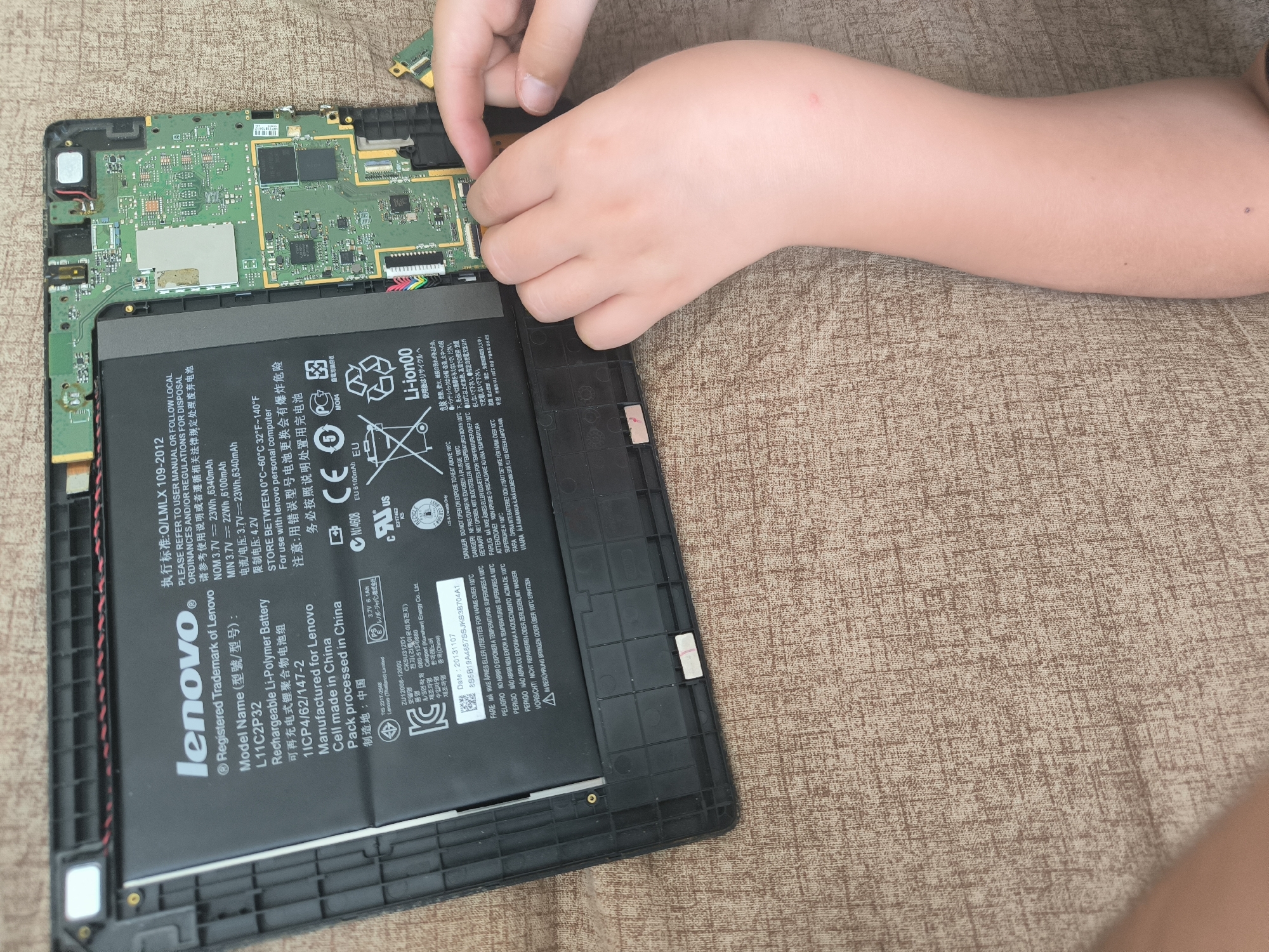 A close-up, overhead shot captures a child's hands working on the internal components of a disassembled Lenovo tablet. The tablet's motherboard, a green circuit board populated with various chips and connectors, is visible in the upper portion of the frame. A large black rectangular battery, labeled "Lenovo," is positioned below the motherboard. The child's fingers are carefully manipulating a ribbon cable connecting to the motherboard, suggesting they are in the process of assembly or disassembly. The tablet is placed on a textured brown fabric surface, resembling a couch or tablecloth. The background is out of focus, with only the brown fabric visible. No specific location in Chișinău, Moldova, is identifiable from the image.