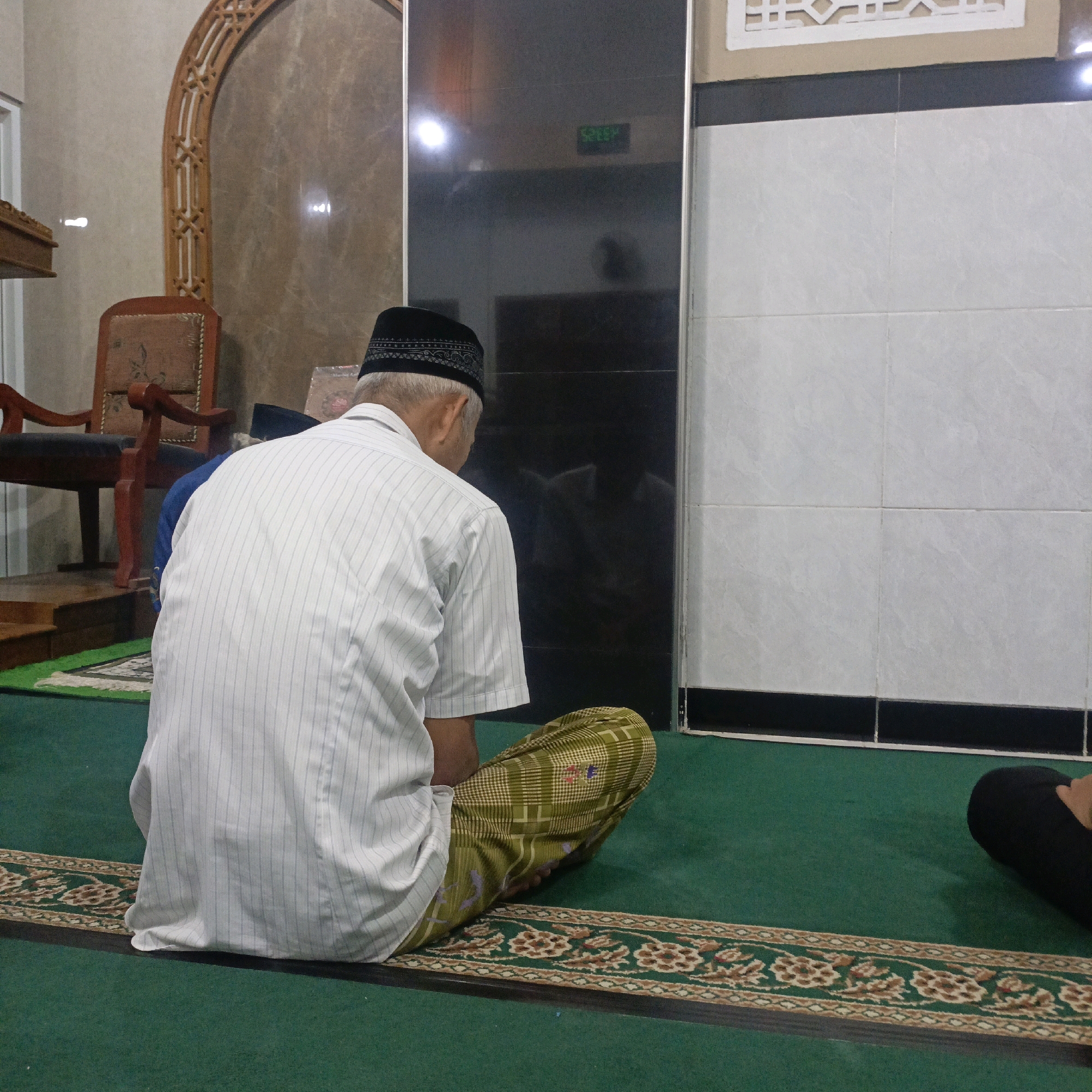 A man wearing a white striped shirt, a black peci, and a patterned sarong is seated on a green carpet in what appears to be a prayer room. He is facing away from the camera, with his head bowed. To his left, a wooden chair with red upholstery is visible. The background includes walls with marble-like tiling and a reflective black surface with a digital display showing "50:08". The carpet has a decorative border with floral patterns. The setting is a prayer room in Salatiga, Indonesia.