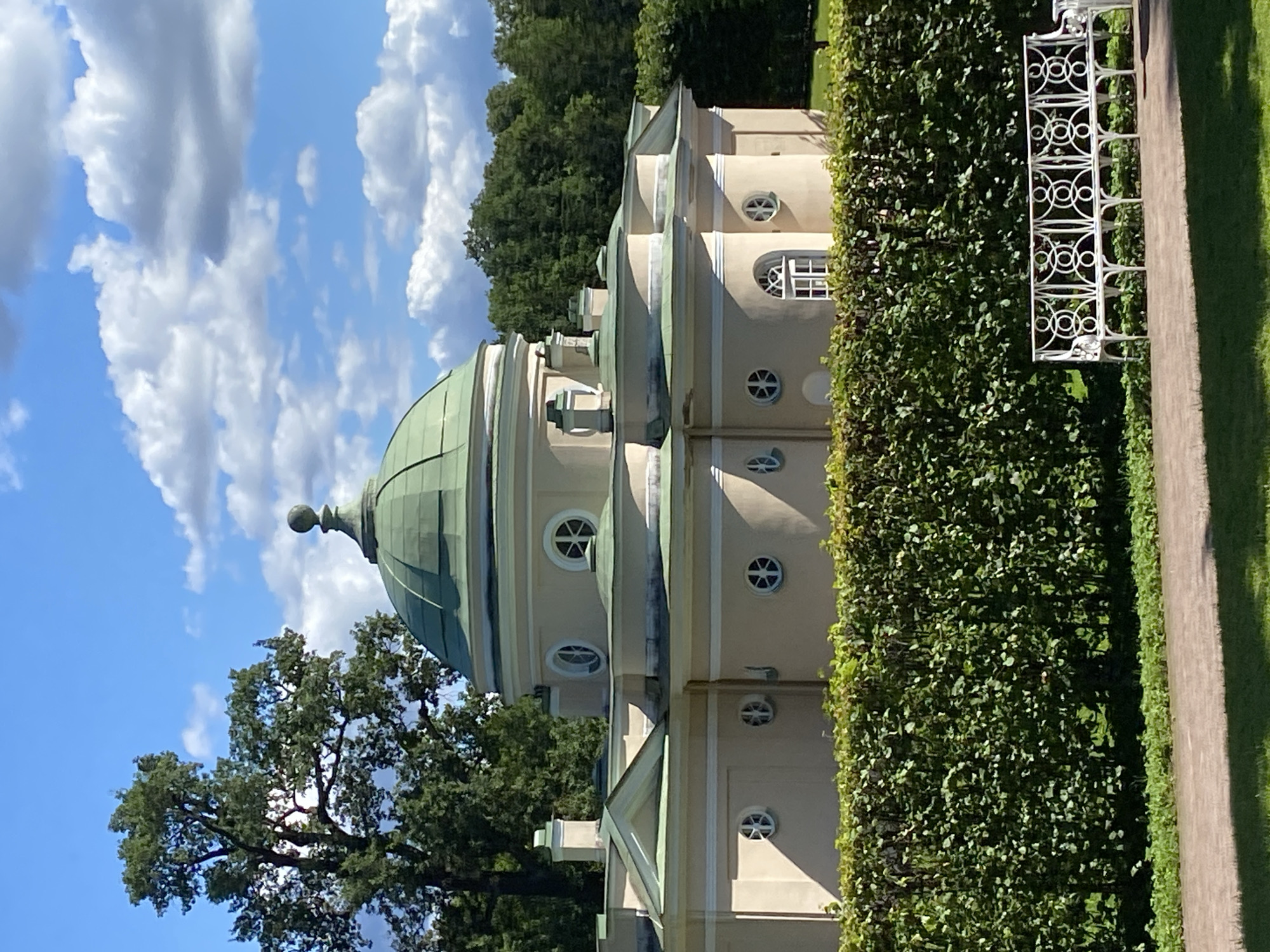 The image shows a view of a pale yellow building with a green domed roof, situated in a park setting. The building has several small, round windows with cross-shaped panes and a larger window with a distinctive arched top and mullioned panes. A neatly trimmed green hedge runs across the foreground, partially obscuring the base of the building. To the right, a white wrought-iron bench sits on a paved walkway. The background consists of lush green trees and a blue sky with scattered white clouds. The architectural style suggests a historical estate. This building is likely part of the Catherine Palace complex in Pushkin, Russia, which is renowned for its grand palaces and extensive parklands. There are no people or animals visible in the scene.