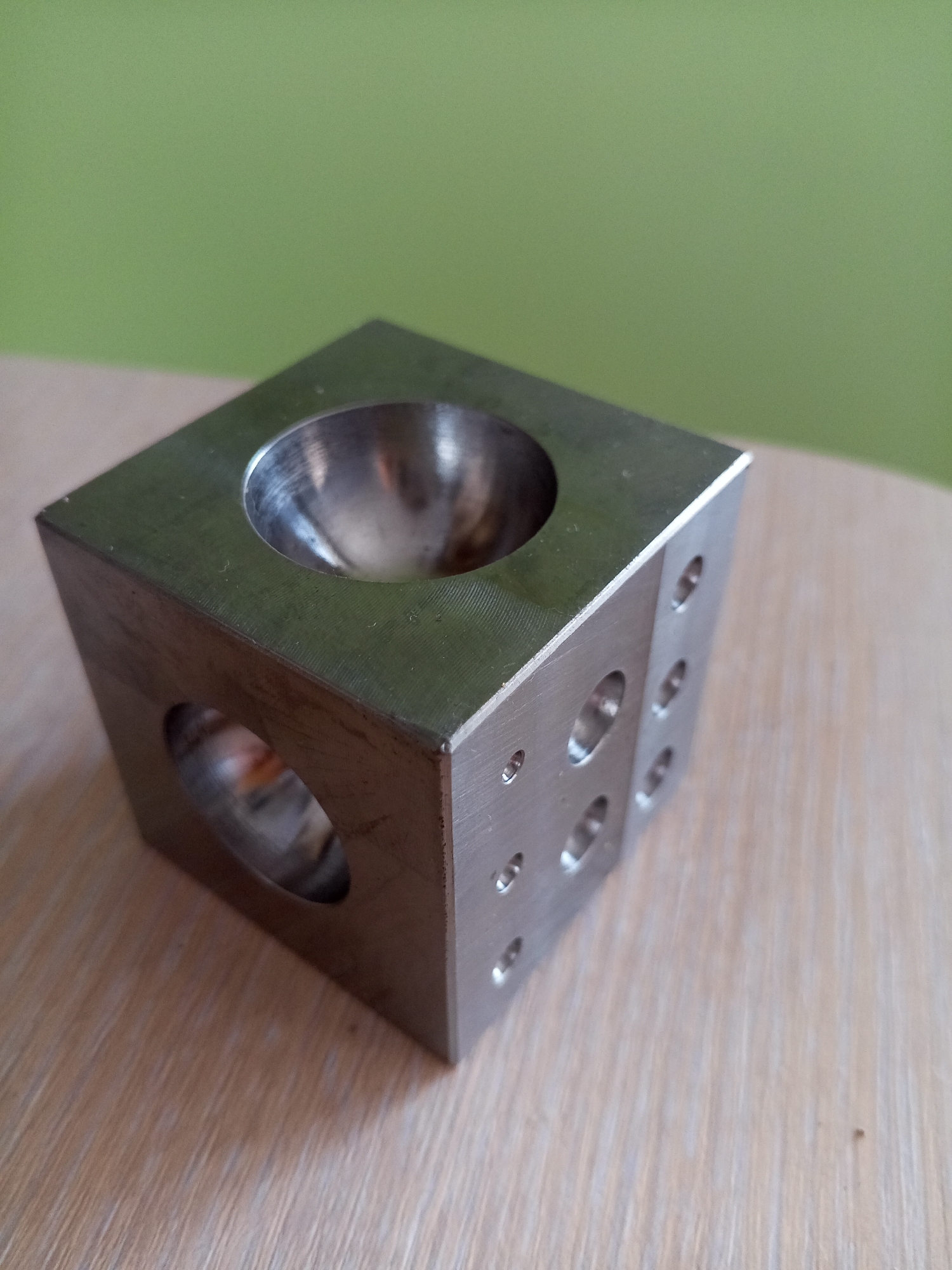 A metallic cube with a concave depression on its top surface sits on a light-colored wooden surface. The cube has several circular holes on its sides, with one large rectangular hole visible on the front face. The background is a solid, soft green. The object is positioned in a dimly lit indoor environment. The image does not contain any elements that suggest it is located in Vilyuysk, Russia.