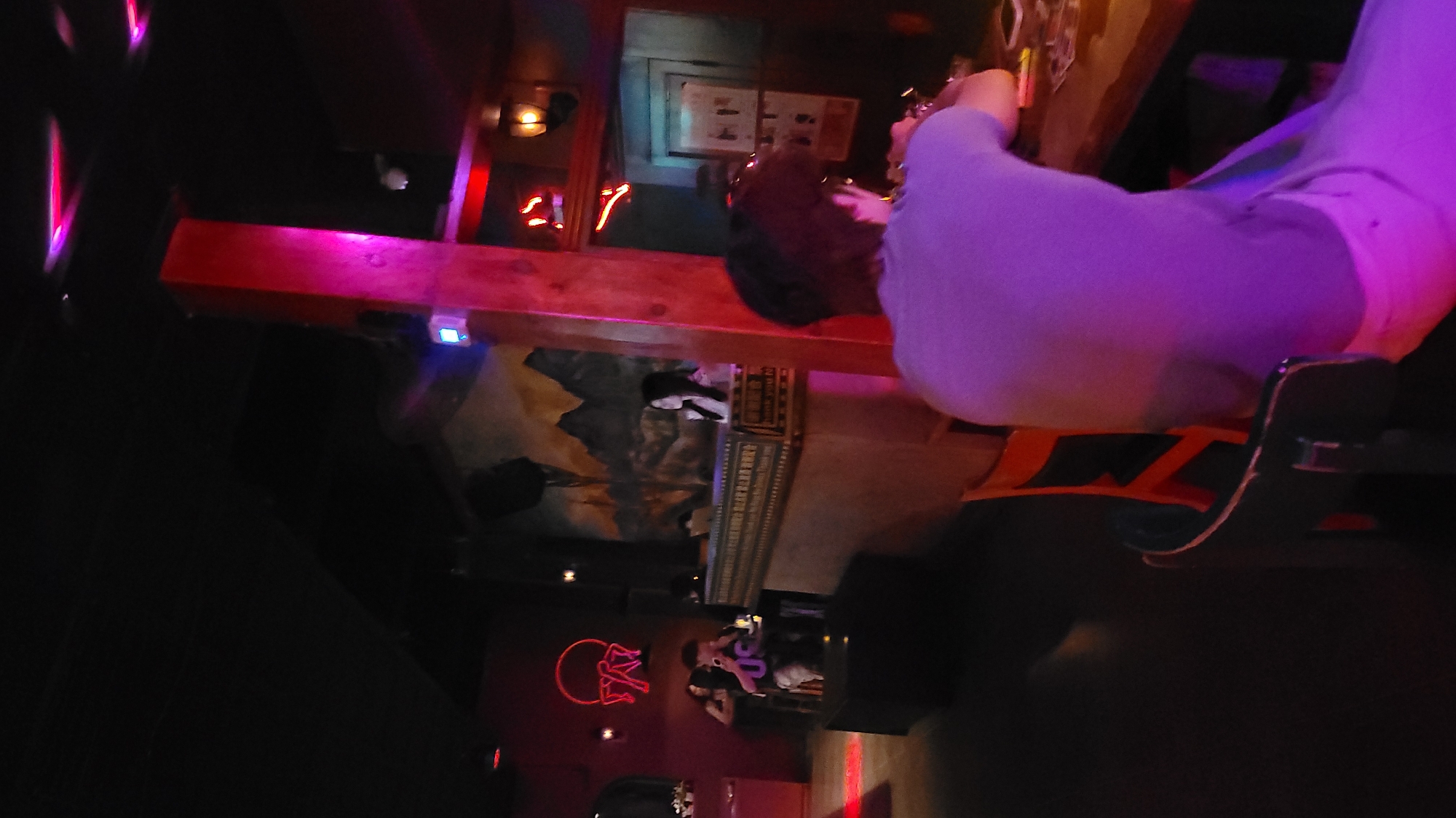 The image captures an indoor scene, likely a bar or pub, at night or in very dim lighting, characterized by vibrant artificial lights. The perspective is from slightly above, looking down into the space.
In the foreground, a person is seated on a bar stool with an orange-colored frame, seen from the back. They are wearing light purple or lavender long-sleeved clothing and are resting their right arm on a prominent wooden bar surface that glows intensely with a strong pink and purple light. The person appears to be holding a glass or drink in their hand.
Behind the illuminated wooden bar, the background reveals a wall decorated with a mural depicting rugged mountains under a sky. Below the mural, shelves hold various bottles or other items, and a vertically oriented box or sign displays the text "LET US ENTERTAIN YOU". Further to the left and deeper in the background, a distinct red neon sign shaped like an animal's head (possibly a bull or ram) glows brightly. Another person is visible in the mid-ground, seated and wearing a dark shirt with light-colored lettering that resembles "09" or "90". A smaller, fragmented red neon light, possibly a number "7", is also visible in the upper left background. A small blue light shines from underneath the main wooden bar.
The overall setting suggests a casual, social atmosphere for an evening out, with the colorful neon lights contributing to a lively ambiance.