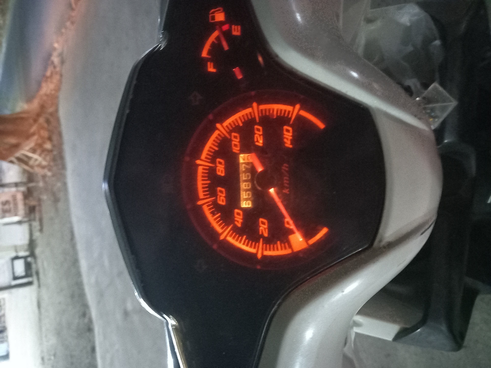This is a close-up, eye-level shot of a motorcycle's instrument cluster. The cluster is illuminated with orange light. The speedometer shows a speed of 0 km/h, and the odometer reads 65873. The fuel gauge indicates the tank is nearly full. The background is blurry and suggests an outdoor setting at dusk or night, possibly on a street in Banyumulek, Indonesia. The surrounding environment appears to be a paved road with some foliage and a building in the distance.