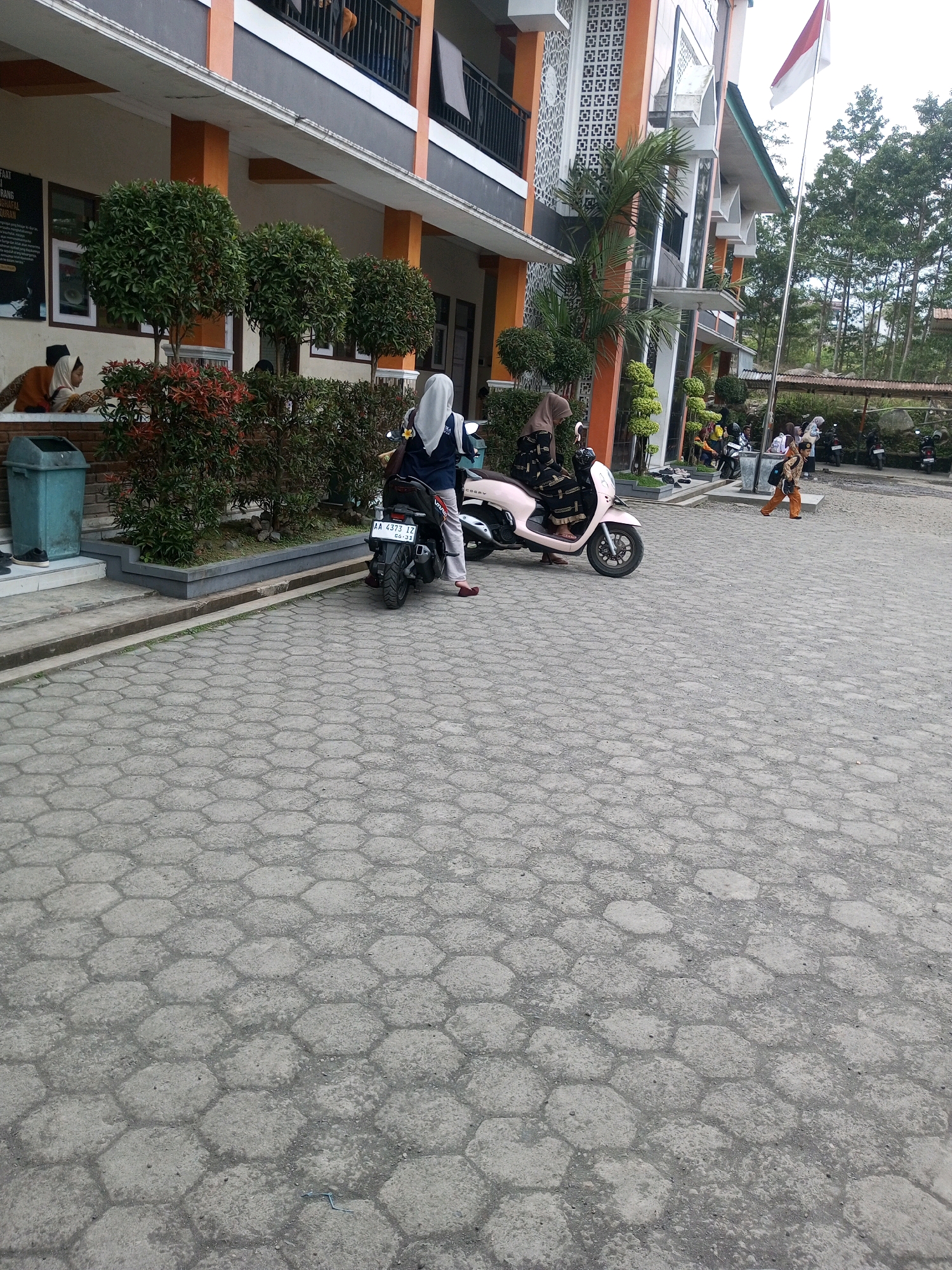 In the city of Kalibeber, Indonesia, an outdoor scene captures a group of individuals near a building with orange and gray accents. A pale pink scooter is parked, with a person in a white hijab and blue top seated on it, facing away from the camera. Next to it, a black scooter is also parked, with a person in a dark hijab and patterned dress standing beside it. A light blue trash can is visible near the building's entrance. The ground is paved with hexagonal concrete blocks. Further in the background, more people are seen near parked motorcycles, and a flag flies from a tall pole. The environment includes lush green trees and foliage, contributing to a slightly overcast atmosphere.