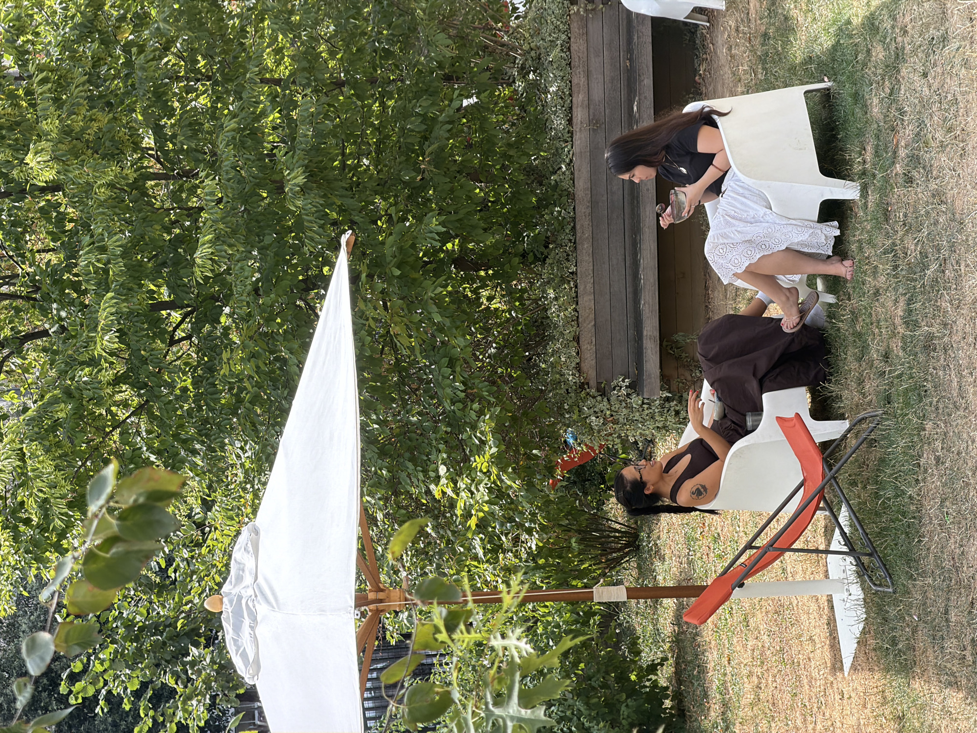 Two women are sitting in chairs under a white umbrella in a garden. The woman on the left is wearing a dark brown dress and glasses, with her hair in a ponytail. She is holding a white cup. The woman on the right is wearing a black t-shirt and a white lace skirt. She is holding a bowl and a spoon. An orange lounge chair is in the foreground. A wooden bench is behind the women. The garden is surrounded by trees and foliage. The image appears to have been taken in Paris, France.