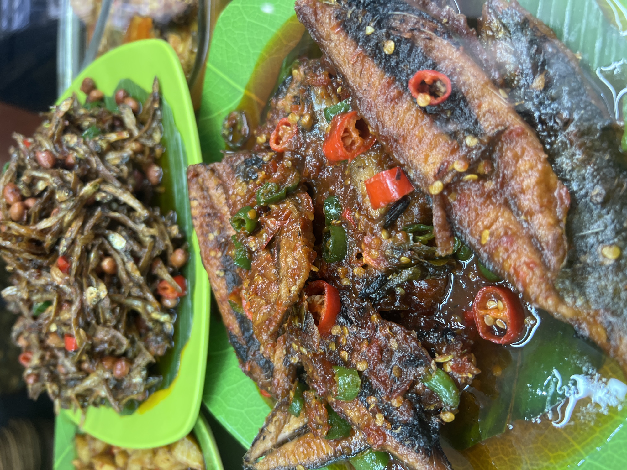 This is a close-up, overhead shot of a dish of fried fish in a spicy sauce, served on a bright green plate with a banana leaf liner, at a food stall in Kuta, Indonesia. The fish, which appears to be small and whole, is coated in a rich, dark red sauce flecked with chopped green chilies and slices of red chili peppers. In the background, another green plate holds a pile of fried anchovies and peanuts, also in a dark sauce. The overall impression is of a vibrant and flavorful Indonesian street food offering.