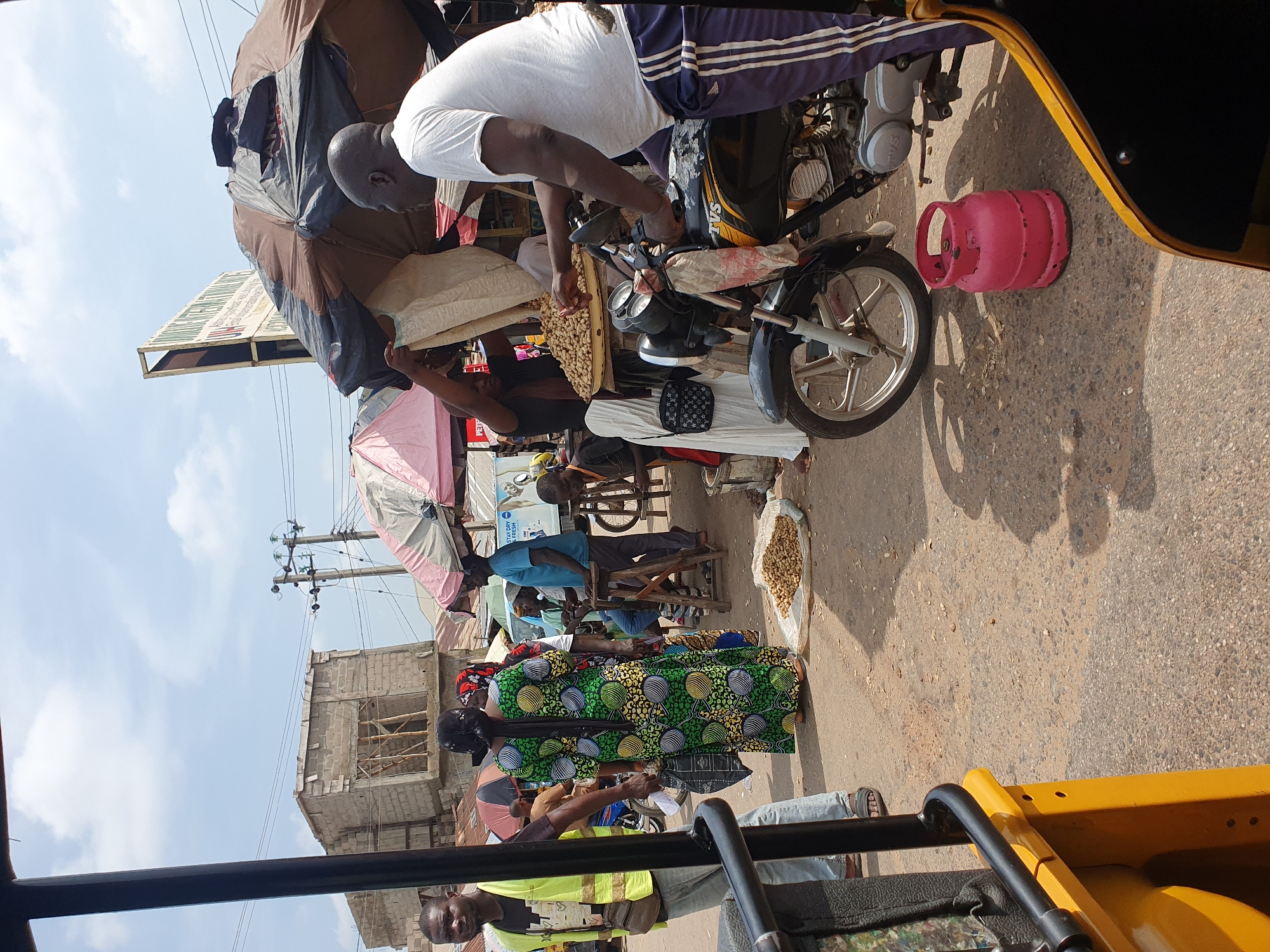 A street market scene in Lokoja, Nigeria, is depicted. In the foreground, a yellow and black vehicle, likely a tricycle or tuk-tuk, is partially visible, suggesting the viewer's perspective from within or alongside it. The ground is a rough asphalt surface.
In the middle ground, several people are engaged in market activities. A man wearing a bright yellow vest and blue jeans stands on the left, looking towards the viewer. To his right, a woman in a vibrant green and black patterned dress is carrying a shopping bag and walking away from the viewer. Several other individuals are visible in the background, some standing and others sitting. One man, wearing a white t-shirt, is standing next to a motorcycle, interacting with a woman who is holding a large sack. A pile of nuts, possibly peanuts, is spread out on a white cloth on the ground. A pink gas cylinder is also placed on the ground near the motorcycle.
Overhead, large umbrellas, some colorful and others dark, provide shade. Buildings, some under construction with visible scaffolding, are in the background, along with utility poles and electrical wires. The sky is partly cloudy. The overall impression is of a busy, open-air market.