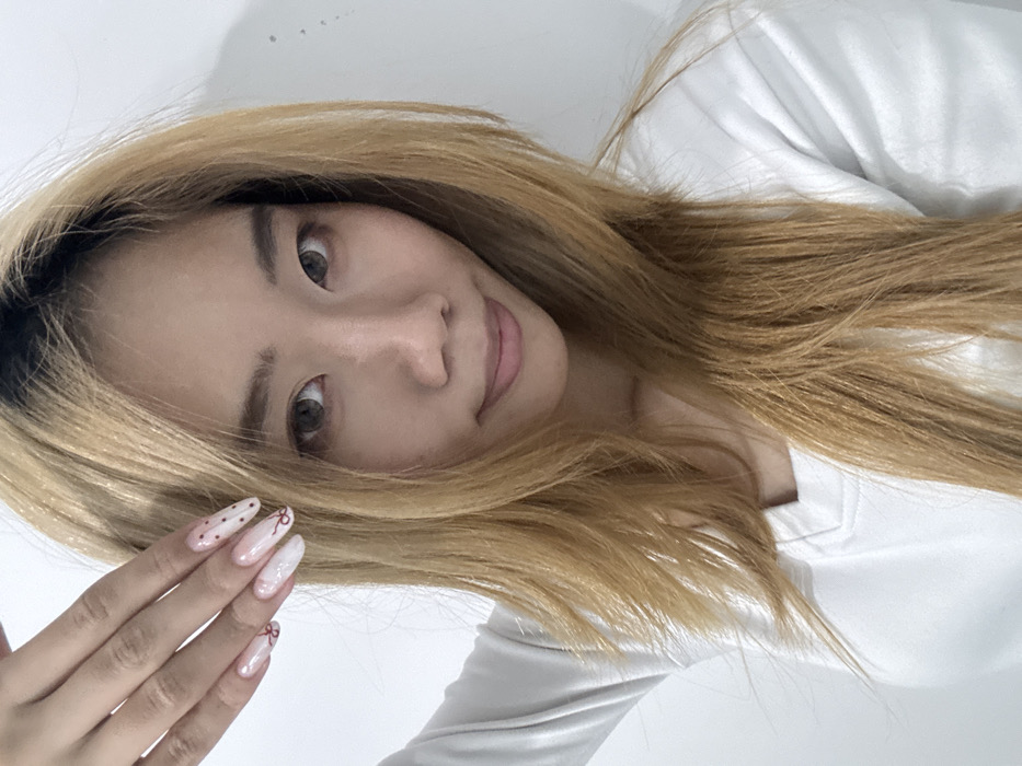A female individual, appearing East Asian, is depicted from the upper torso upwards against a plain white wall. The indoor setting lacks specific visual identifiers for a precise location within Yuzhong District, China. She has long, layered, light brown hair with darker roots and wears a white, V-neck, long-sleeved top. Her eyes are dark, appearing to utilize grey-hued contact lenses, and her expression is a slight smile directed at the camera. Her left hand is raised to shoulder level, displaying long, almond-shaped fingernails. These nails exhibit an ombré design with white tips fading into a translucent base. Embellishments include small reddish-brown bow designs on some nails and reddish-brown polka dots on others.