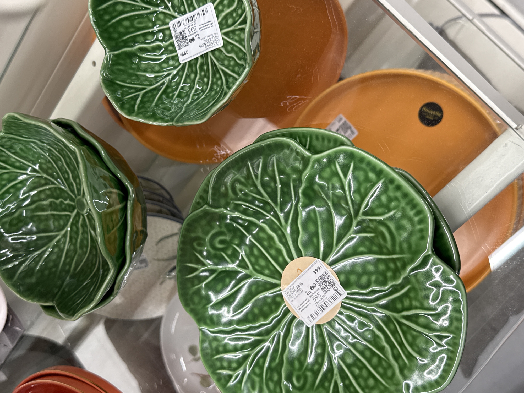 The image shows a display of decorative tableware, primarily featuring green cabbage leaf-shaped plates and orange circular plates. The green plates are stacked and have price tags with Russian text indicating a discount and price, identifying the brand "TH ROYAL UNION" and "SONO A CASA". The price tags also contain QR codes. Some orange plates have a "Nouvelle" brand label. The items appear to be on shelves within a store, possibly in a city environment. The text on the labels indicates a location somewhere that uses the Russian language, such as the city of Izhevsk, Russia.
