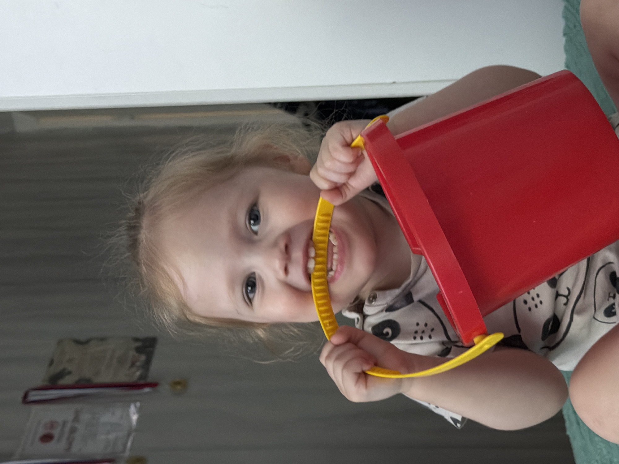 An indoor, frontal image, taken in the city of Perm, Russia, depicts a young child, likely a toddler, with light-colored hair and blue eyes, smiling broadly and looking directly at the camera. The child is wearing a white short-sleeved garment patterned with black animal faces. They are holding a red plastic bucket with a yellow handle, actively biting the ribbed section of the handle with their front teeth. The background to the left features a muted gray wall with three hanging items, appearing to be framed certificates or awards, two with visible red ribbon accents and one with a gold medal. To the right, a vertical white architectural element, consistent with a door frame or wall, is visible. The child is seated on a textured surface of a greenish-blue color.