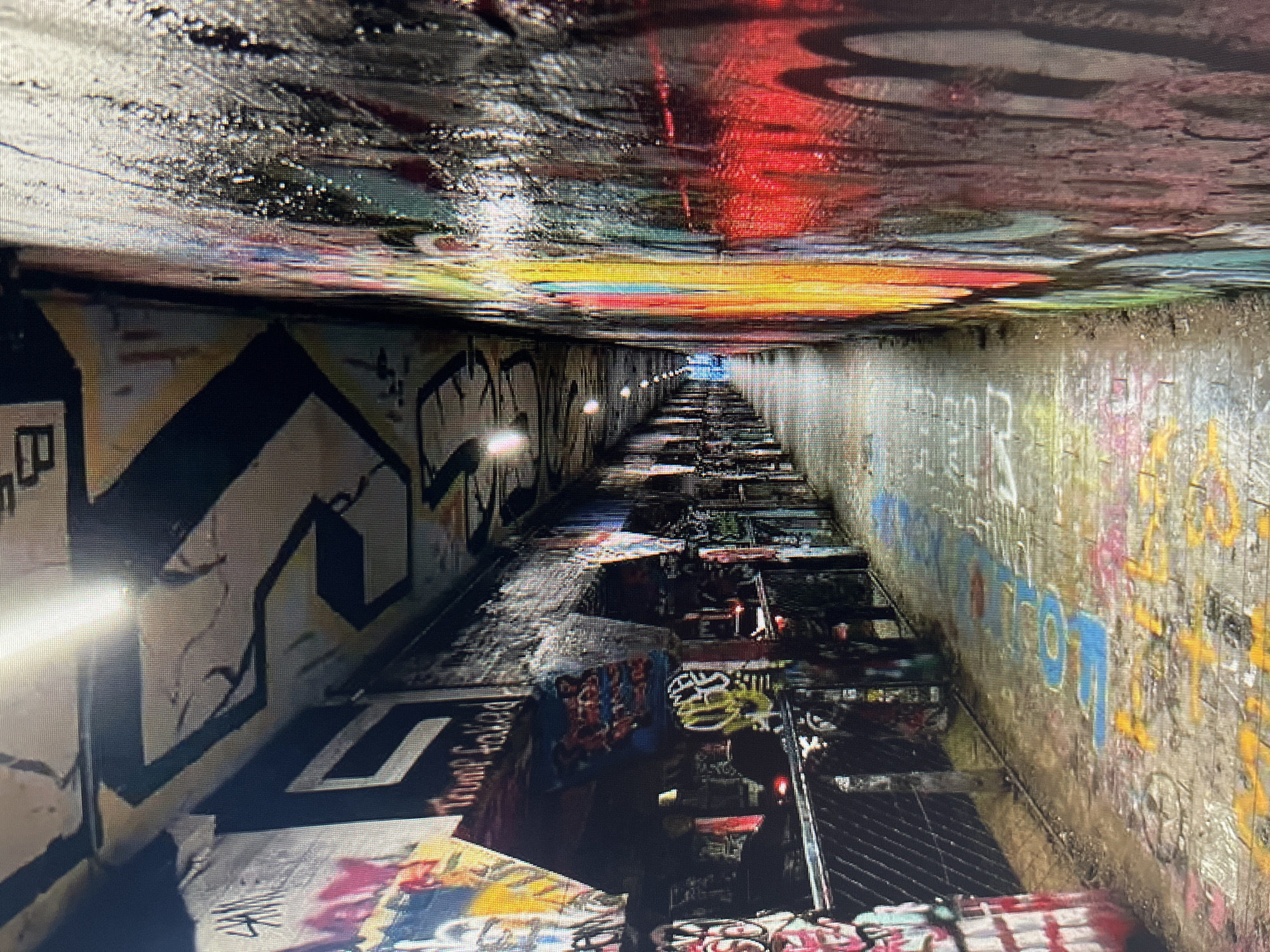 The image depicts a graffiti-covered tunnel or underpass, possibly a pedestrian walkway, in an urban environment. The walls and ceiling of the tunnel are heavily adorned with spray-painted graffiti in a variety of colors and styles. The floor of the tunnel is also covered with graffiti. The lighting in the tunnel is provided by overhead lamps, casting a dim glow on the scene. The perspective suggests a long, narrow passageway extending into the distance. There are no people or specific identifying landmarks visible in the image that would pinpoint the exact location. The overall impression is one of urban decay and artistic expression through street art.