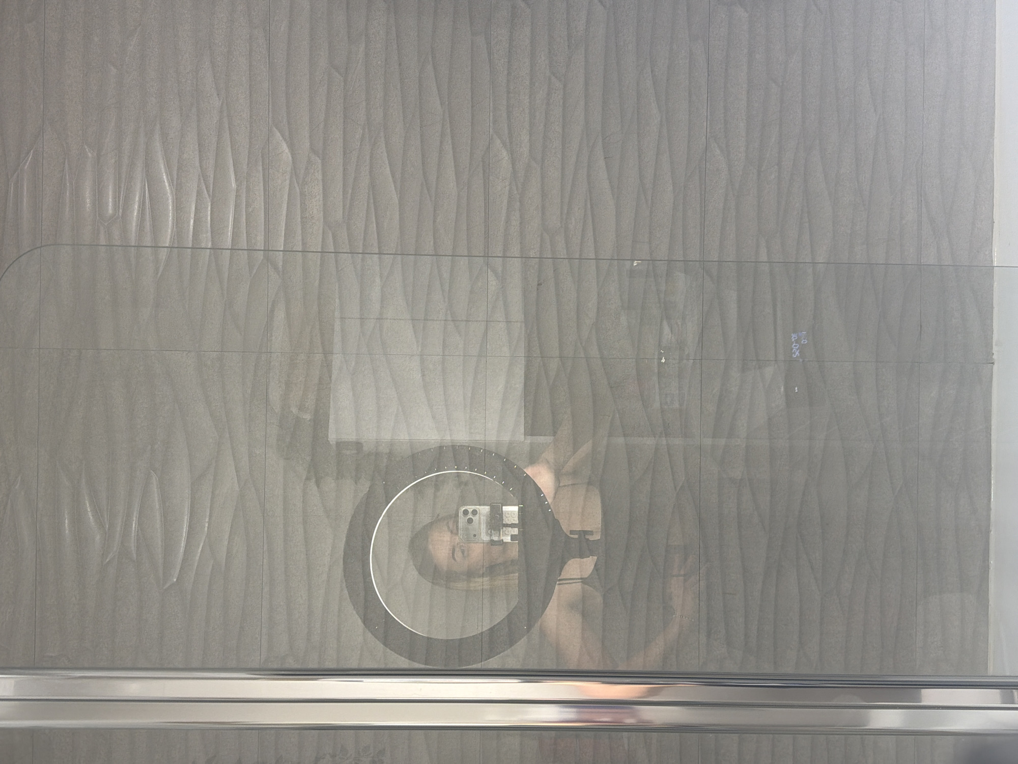 The image displays a selfie being taken through the reflection in a clear glass panel, set against a textured gray tiled wall. The main subject is a person, partially visible in reflection, holding a ring light with a smartphone mounted in its center. The person's face and upper body are reflected, looking towards the smartphone's camera.
The scene is an indoor setting, possibly a modern bathroom or shower enclosure, indicated by the tiled wall and the clear glass panel with a metallic rail at the bottom. The background consists of gray tiles with a distinct wavy or rippled texture.
The activity taking place is a self-portrait (selfie) or video recording, evidenced by the use of a ring light for illumination and the smartphone pointed at the person. On the reflected smartphone screen, faint digital text or numbers, possibly "30:00:05," can be observed, suggesting a timer or recording duration. The person's expression appears neutral or slightly smiling.
Notable details include the bright, circular illumination from the ring light, which highlights the person's face. The overall lighting suggests an indoor environment, likely independent of natural daylight. The image is set in Sofiivska Borshchahivka, Ukraine.