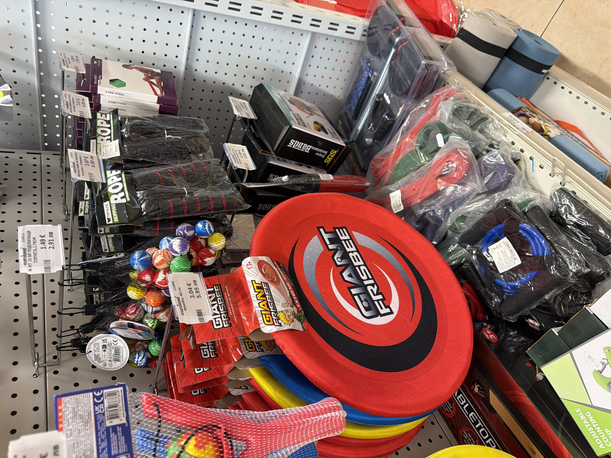 The image displays a retail store shelf stocked with various exercise and outdoor toys. In the foreground, a large red "GIANT FRISBEE" is prominently displayed, along with several other frisbees in different colors. Behind the frisbees, there are bags of colorful small balls, skipping ropes in black packaging, and yoga belts. Further down the shelf, exercise resistance bands and yoga mats are visible. The products are arranged on metal shelves against a pegboard wall. Price tags and product labels are attached to many of the items. The store appears to be a general merchandise or sporting goods store. The presence of Bulgarian currency and labeling suggests the location is in Bulgaria. Given the provided context that the image is from Pazardzhik, Bulgaria, this scene depicts a retail environment within that city. There are no people visible in the image.