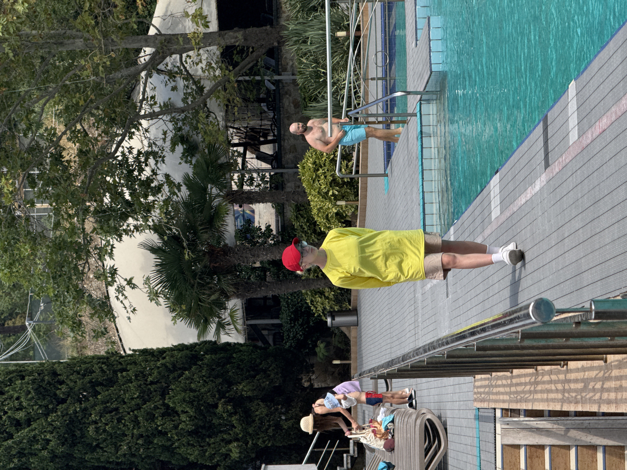 In Массандровский поселковый совет, Ukraine, a man with a bald head and beard, wearing blue swim trunks, stands at the edge of a bright turquoise swimming pool. He is partially submerged in the water, with his hands resting on a silver-colored metal railing that surrounds the pool. To the left of the pool, a woman wearing a straw hat and a dark top is helping a young child, who is wearing red swim trunks and a blue hat, to put on a white shirt. Behind them are lounge chairs and a large tree. In the foreground, a person in a bright yellow long-sleeved shirt and khaki shorts, wearing a red cap and glasses, walks away from the camera along the pool deck, which is made of gray composite planks. The background features lush green trees, palm trees, and a white tent-like structure.