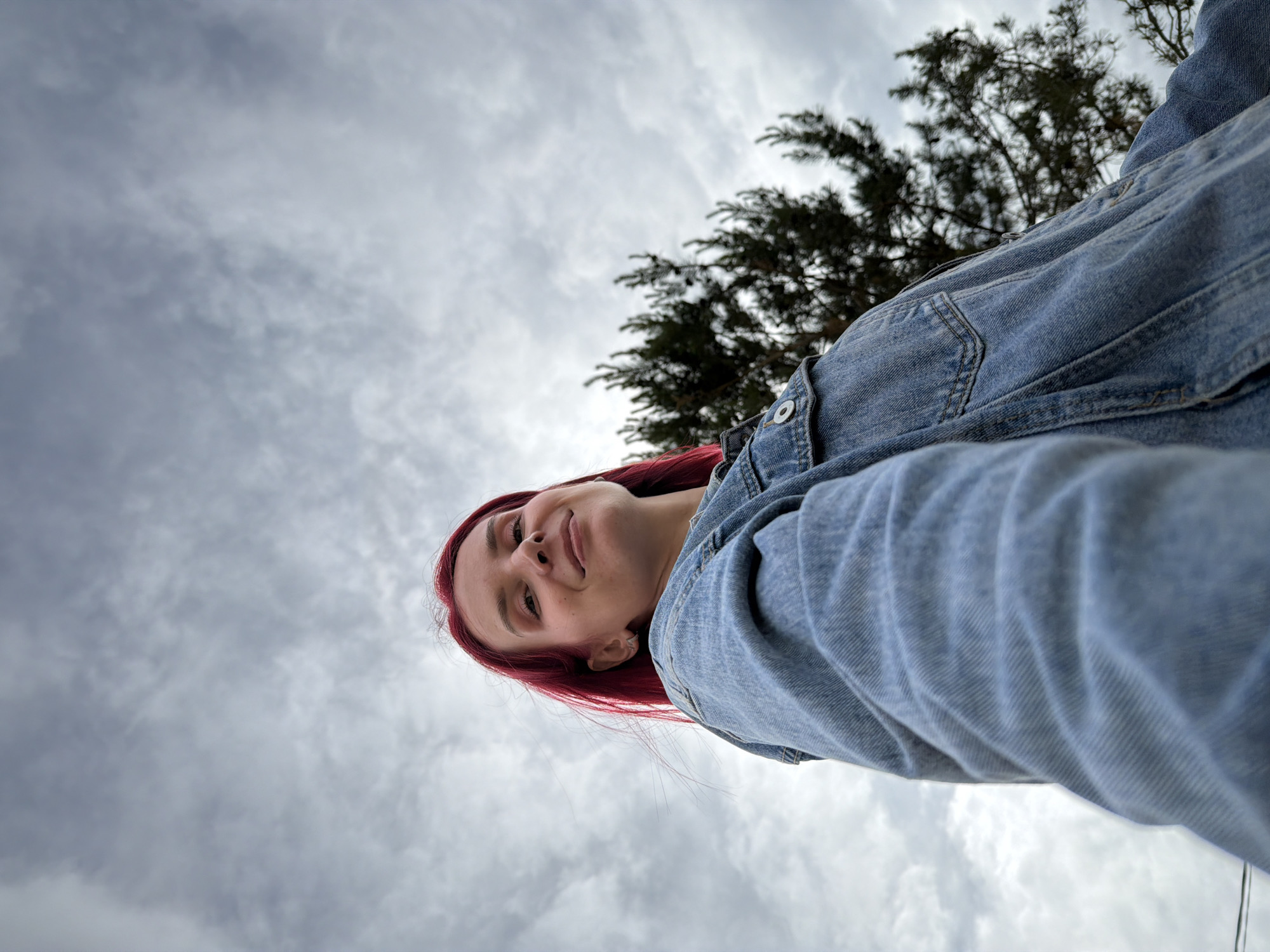 An outdoor, low-angle shot captures a young woman with long, vibrant red hair against a cloudy sky. The image is tilted, presenting the woman diagonally across the frame, with her head towards the lower left and her torso extending towards the upper right. She is wearing a light blue denim jacket, with its texture and stitching clearly visible. Her face is turned slightly upwards and to her right, featuring a calm expression with a subtle, gentle smile. Her eyes are open, looking towards the camera's perspective.
The background is dominated by an overcast sky, filled with soft, diffused grey and white clouds, indicating daytime. In the upper right portion of the frame, behind the woman, a portion of a dark-leaved tree is visible, adding a natural element to the setting. The low perspective suggests the camera was placed below the woman, looking up at her. There is no visible text in the image. The setting is outdoors, possibly in a park or open area in Chaikovsky, Russia, characterized by the natural light and foliage.