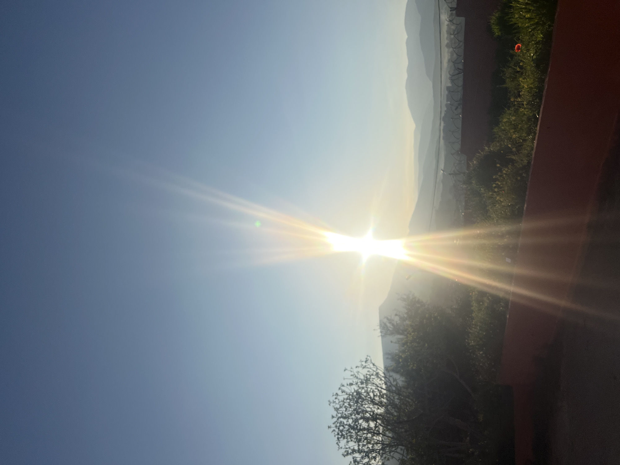 The image captures a sunrise over a landscape in Ouaouizert, Morocco. The sun is prominently positioned in the center, emitting bright rays. Mountains are visible in the distance, partially obscured by the glare of the sun. A barbed-wire fence runs along the upper right side, bordering vegetation. A red structure is on the right edge of the image, possibly part of a building or wall. In the lower right corner, there is vegetation. The sky is a clear blue color, becoming lighter near the horizon.