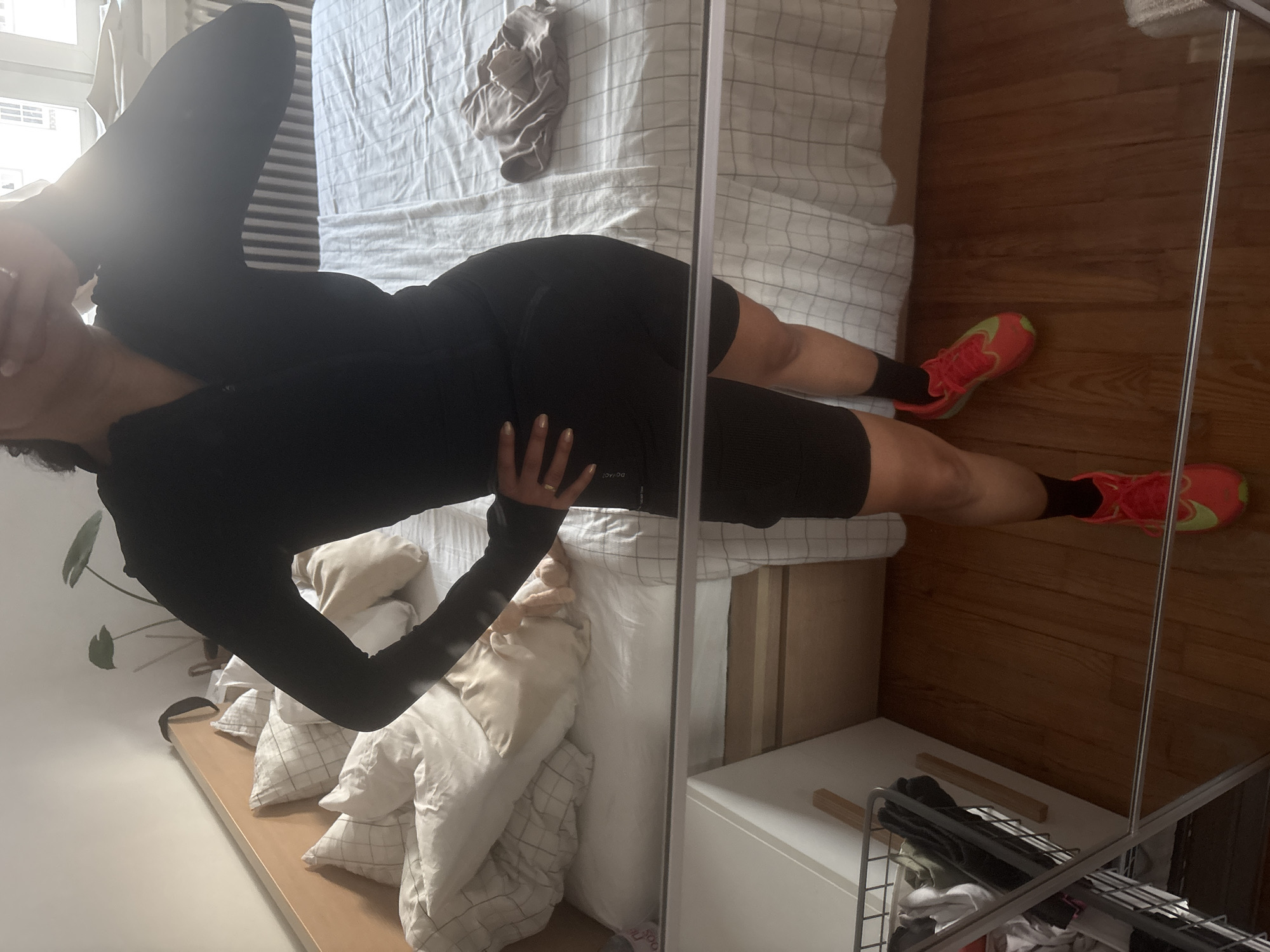 The image depicts an indoor scene, likely a bedroom, viewed from a slightly elevated angle. The primary subject is a person, appearing female, lying on their back on a bed, with their head to the left and feet to the right of the frame. Their left arm is bent with the hand partially covering their face, while their right hand rests on their upper thigh. The person wears a long-sleeved black top and black cycling shorts. On their feet are vibrant red-orange athletic shoes with lime green accents and black socks. A gold-toned ring is visible on their left hand, which also has light-colored nail polish.
The bed is covered with white and grey checkered bedding, and several pillows—some white, some beige, and some matching checkered—are piled at the head of the bed. A crumpled light brown piece of fabric lies on the bed to the person's right.
To the right of the bed, a polished wooden floor is visible. A tall, slender mirror frame extends vertically through the image, creating a reflection of the room. In the background on the far left, a bright window with horizontal blinds suggests natural daylight. A green plant is partially visible near the head of the bed. Below the foot of the bed, a white bedside table or chest of drawers is present, topped with a metal wire basket containing folded clothes. Faint text, "DOYAD," is visible on the person's left short leg. The scene captures a moment of the person posing in a relaxed manner.